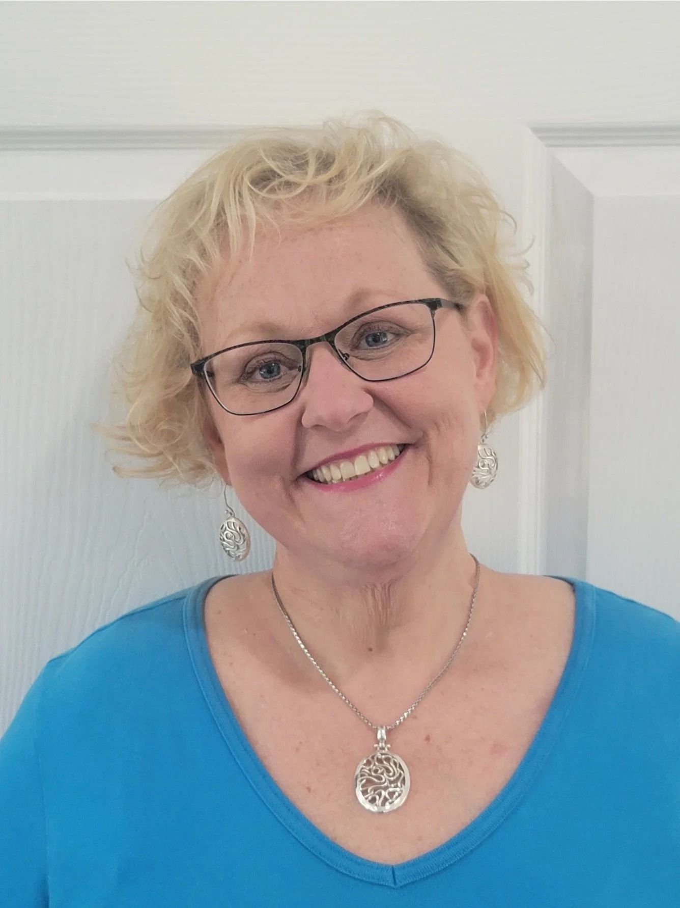 Woman with blonde curly hair, glasses, smiling, wearing a blue shirt and silver jewelry, in front of a white door.