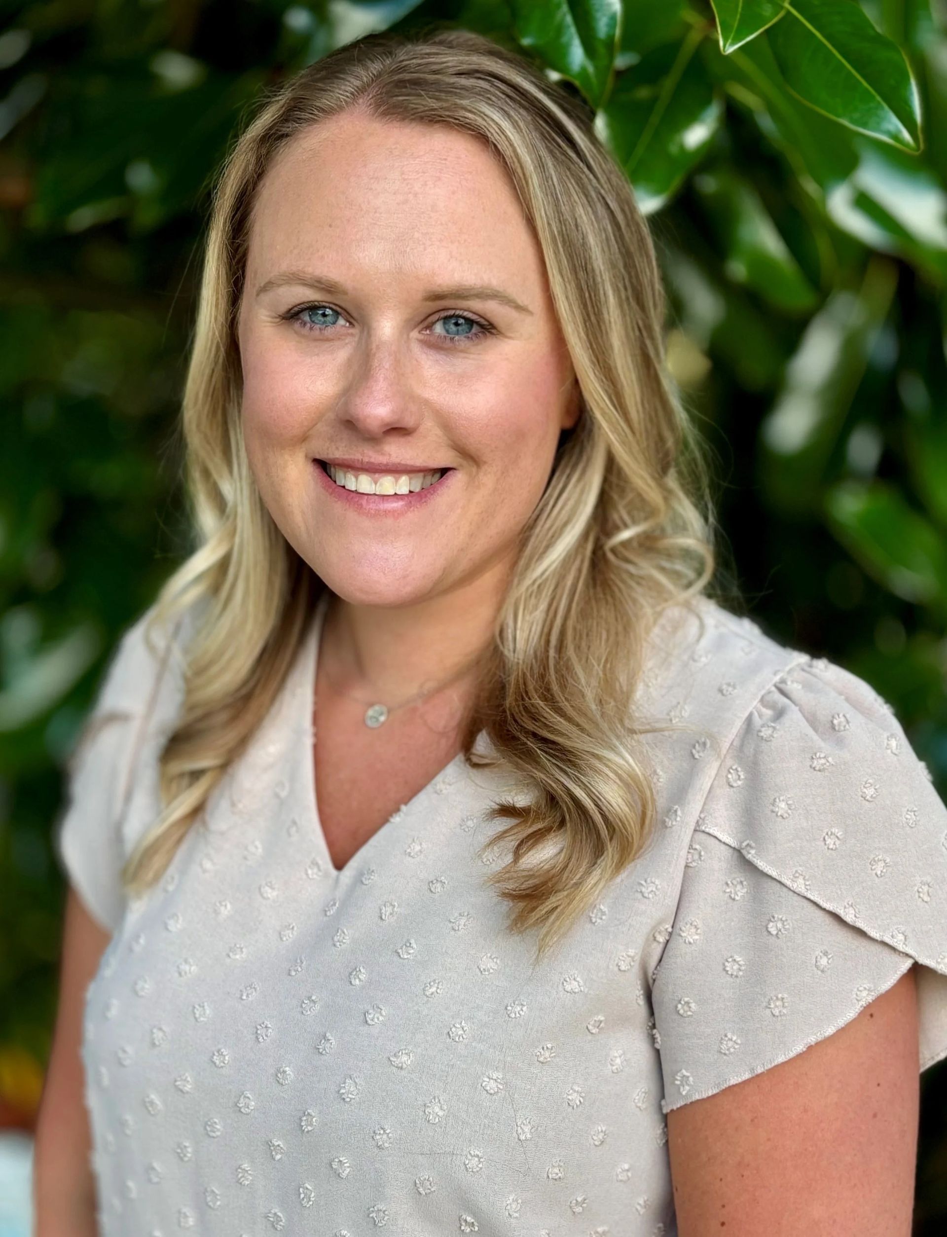 Blonde woman smiling, wearing a light-colored blouse, in front of leafy green background.