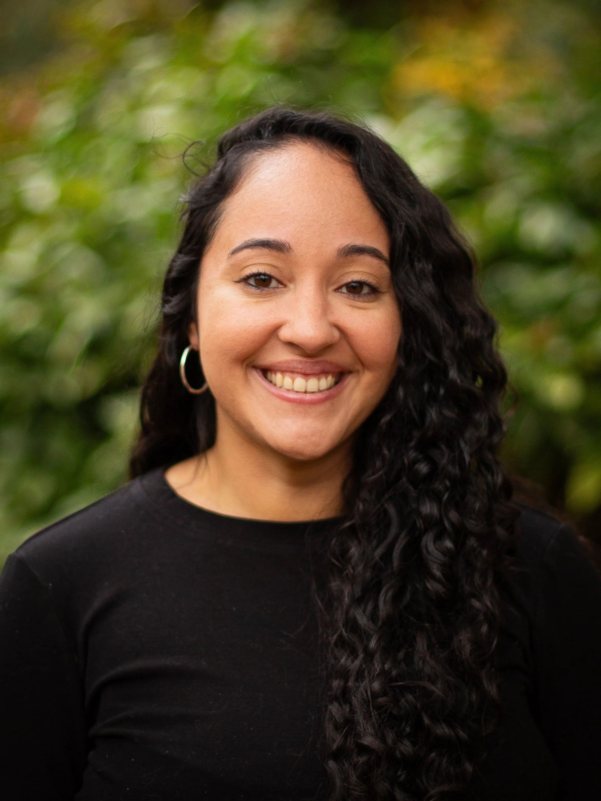 Woman with curly dark hair smiles, wearing a black shirt, silver hoop earrings, set against a green, blurred background.