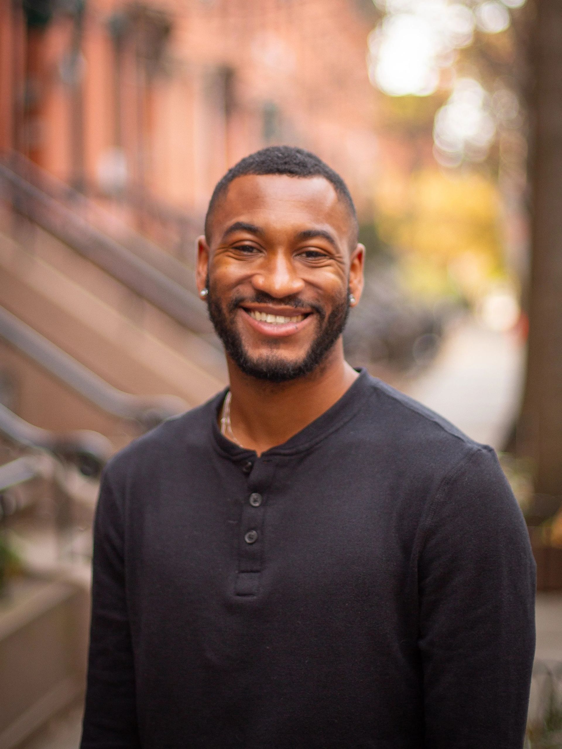 Man with a beard smiles, wearing a black shirt, standing outside a brick building.