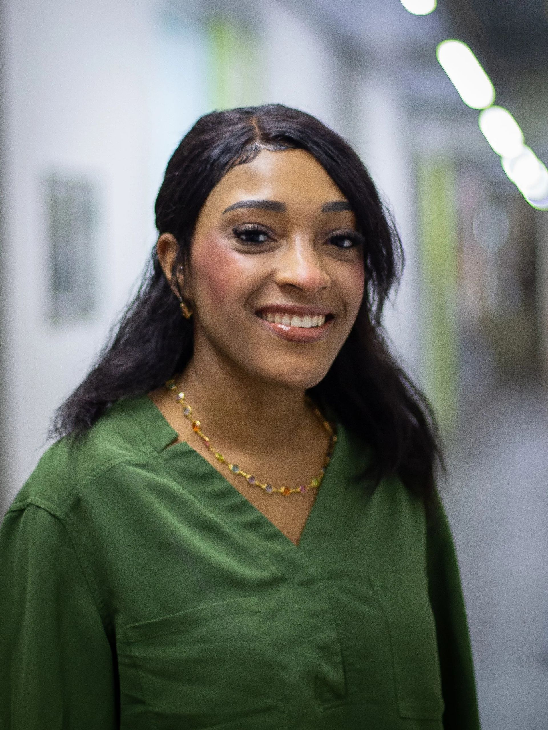 Woman with dark hair smiles, wearing a green top, gold necklace, in a hallway.