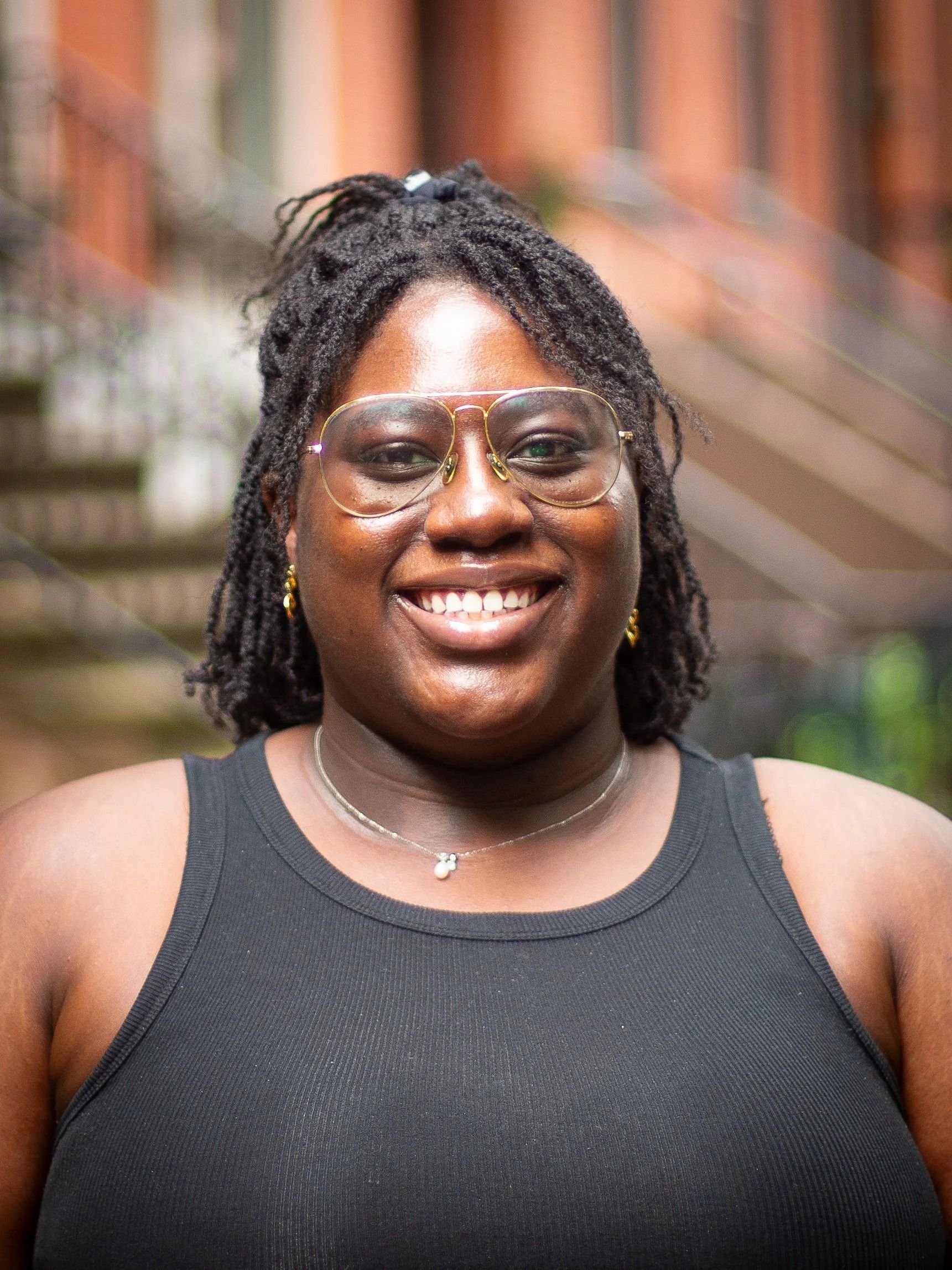 Woman with locs, clear glasses, smiling in front of a brick building. She's wearing a black tank top and necklace.