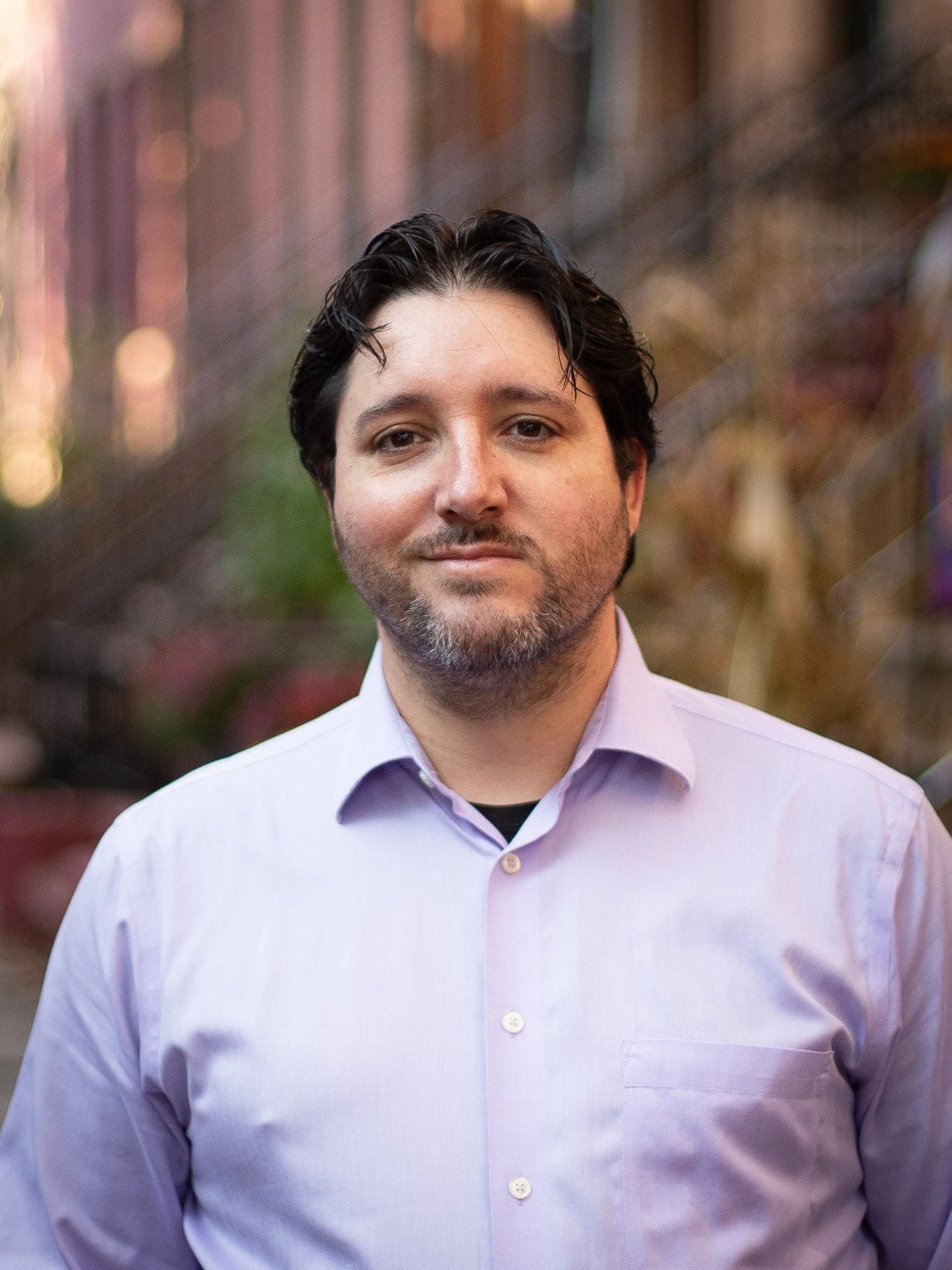 Man in light purple shirt smiles at camera, against a blurred outdoor background.