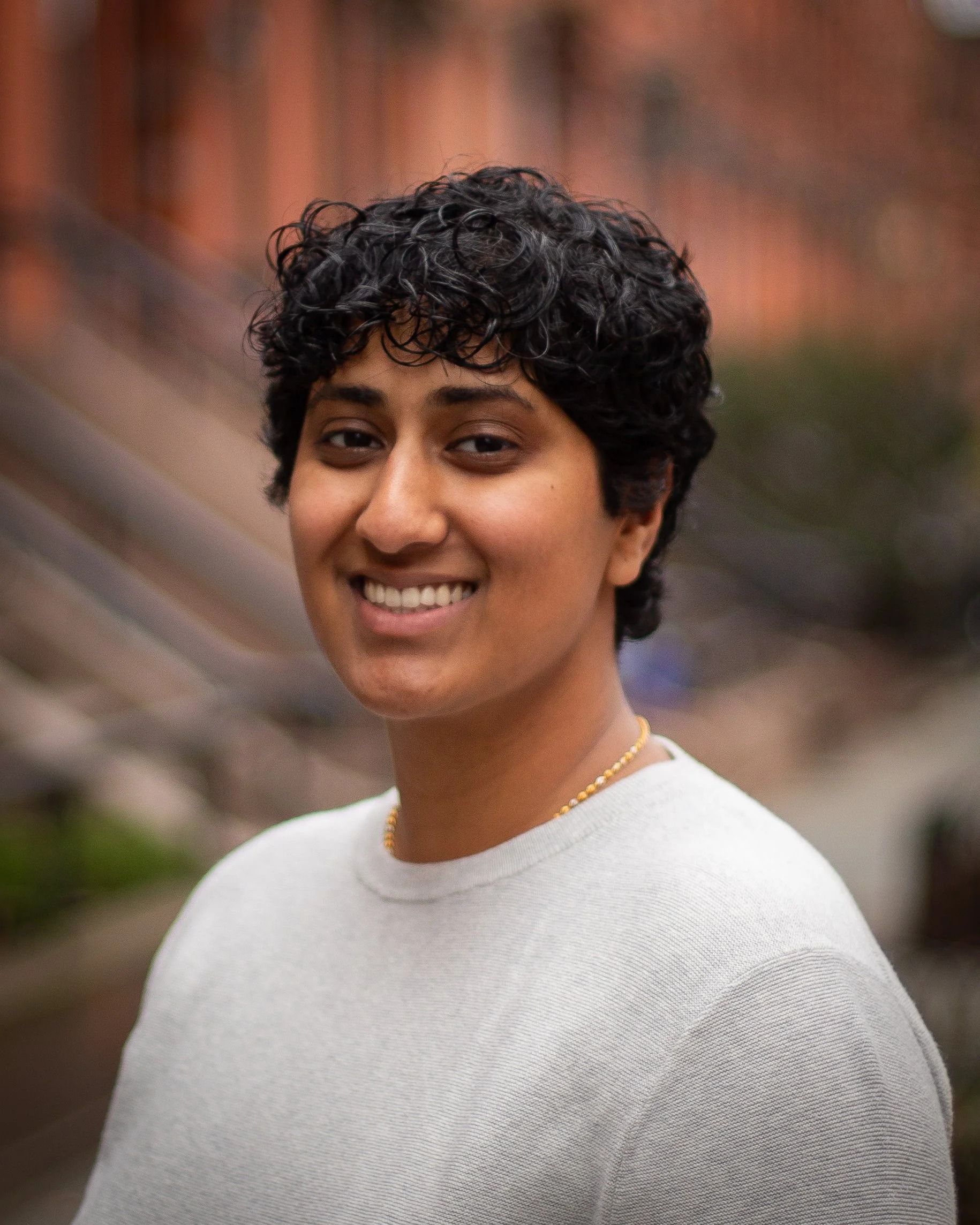 Person smiling, wearing a light gray sweater, standing outdoors with a blurred brick background.