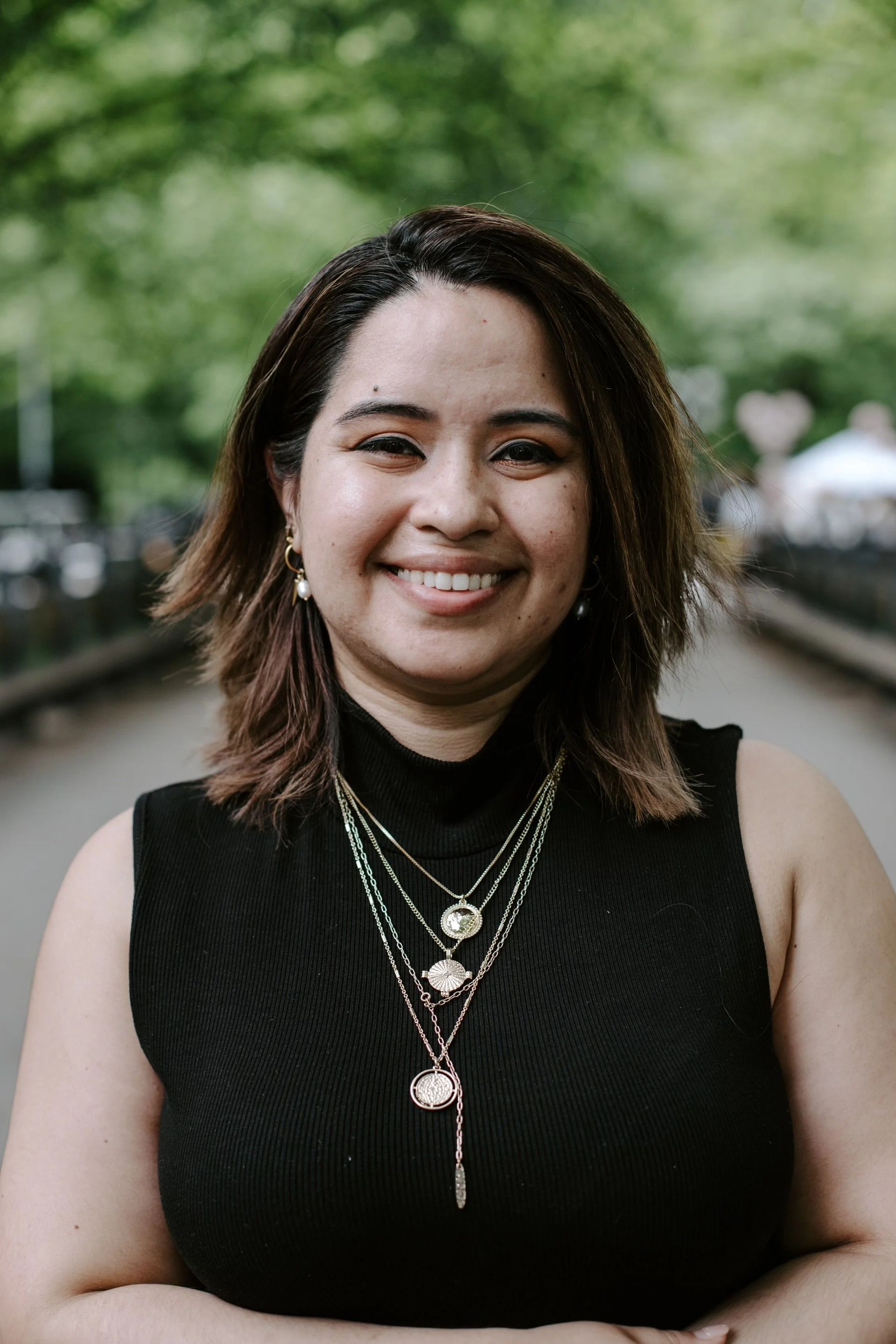 Woman with short brown hair smiles, wearing black top and multiple necklaces, outdoors on a bridge.