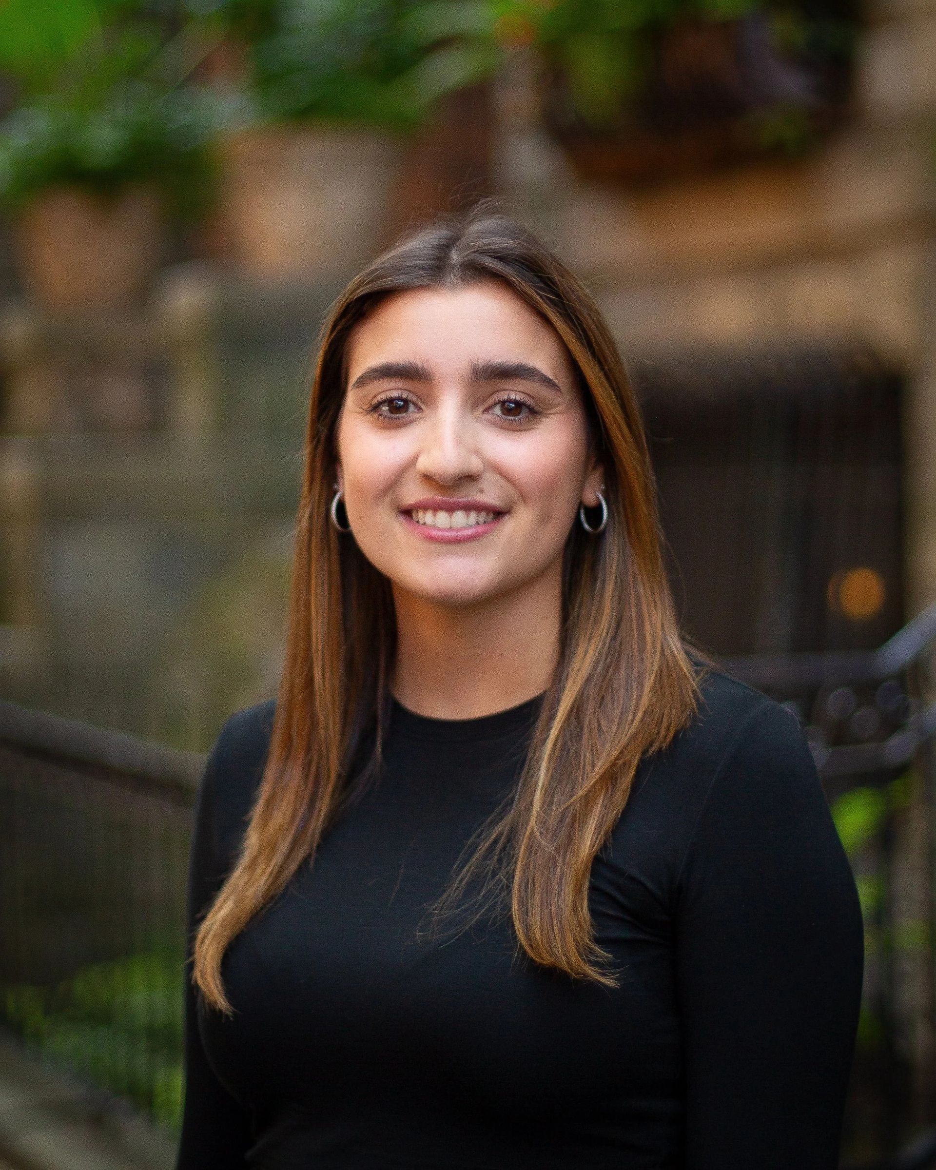 Woman in a black shirt smiles outdoors, brown hair, silver hoop earrings, blurred background.