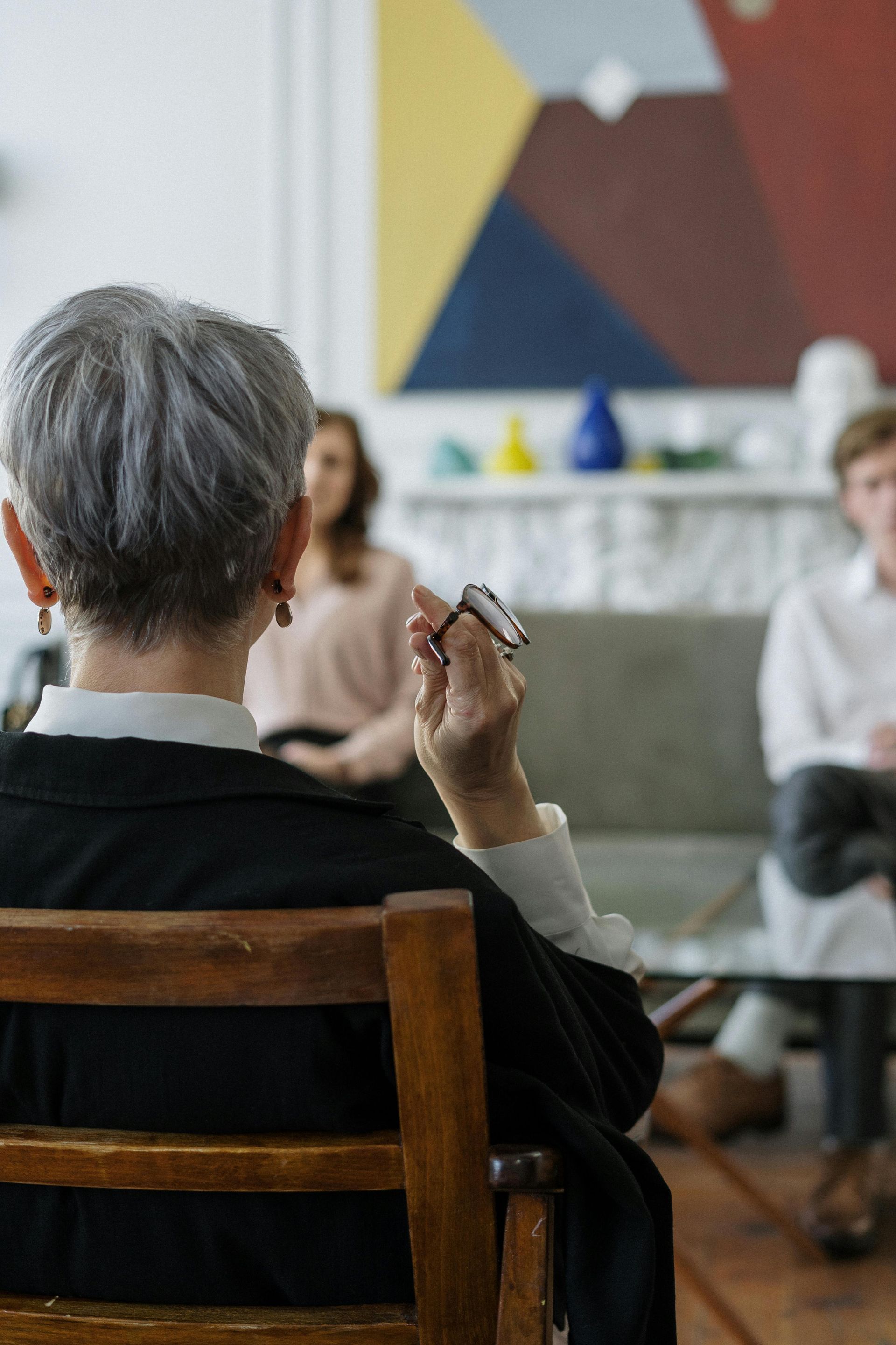 Therapist in a chair, facing two people on a sofa, holding a pen, abstract art on the wall.