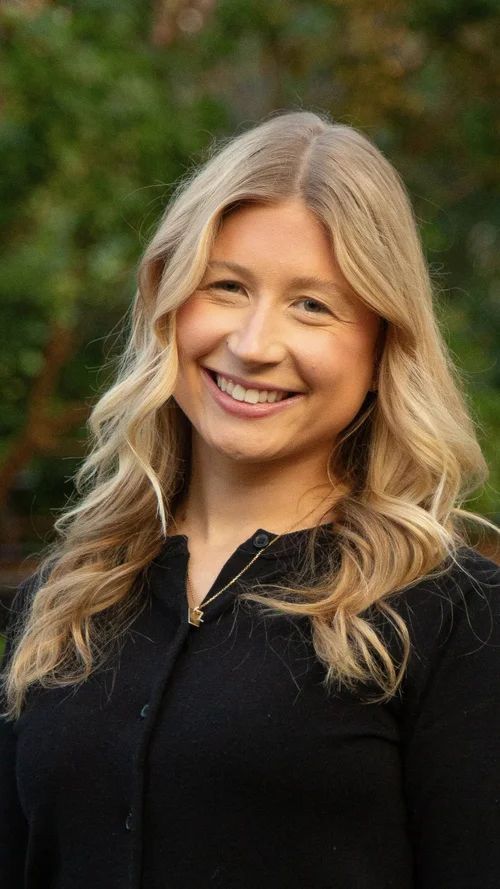 Woman with blonde hair smiling outdoors, wearing a black top.