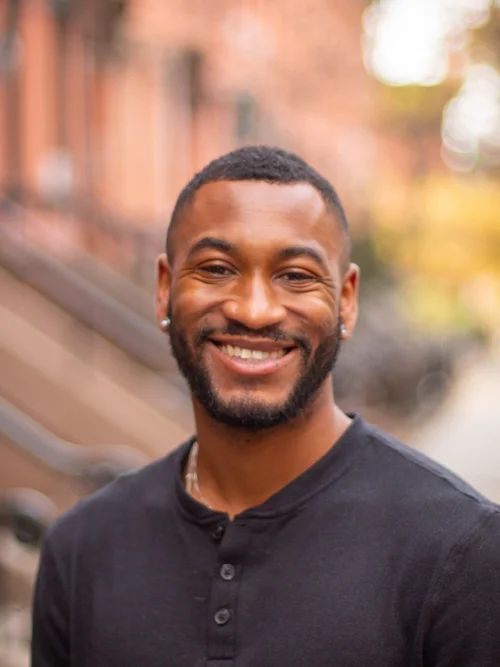 Man with short dark hair and beard smiles broadly outdoors, brownstone background.