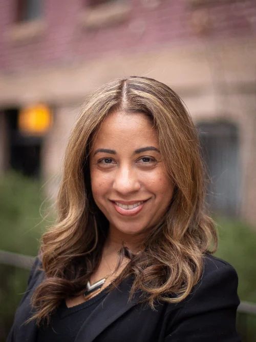 Woman with long wavy hair smiles, wearing a black blazer. Building background.