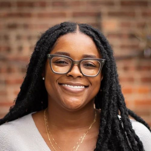 Woman with glasses smiles, looking at camera. Long black locs, light-colored top, and brick background.