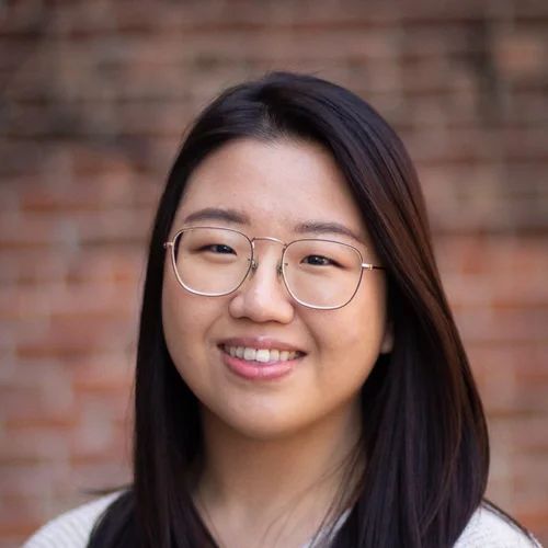 Woman with glasses smiling in front of a brick wall.