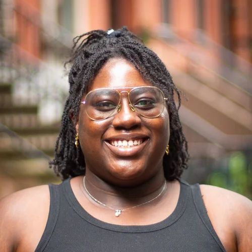 Person with glasses smiling outdoors, wearing a black tank top, and jewelry.