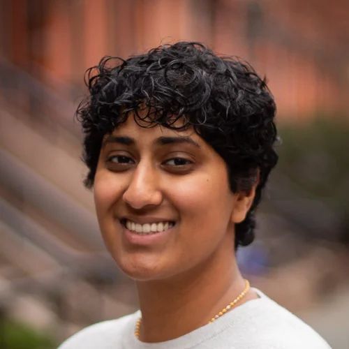 Woman with curly dark hair smiles outdoors, wearing a light shirt and necklace.