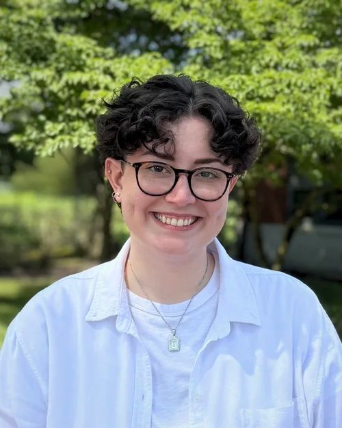 Person with curly dark hair wearing glasses and a white shirt smiling outdoors in front of greenery.