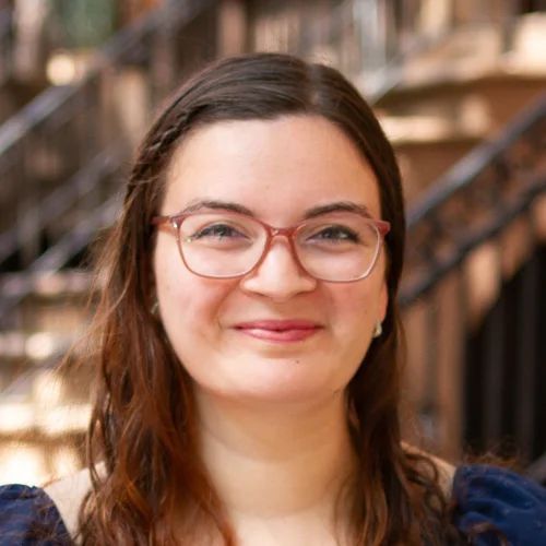 Woman with glasses smiles at the camera in front of a brick building with stairs.