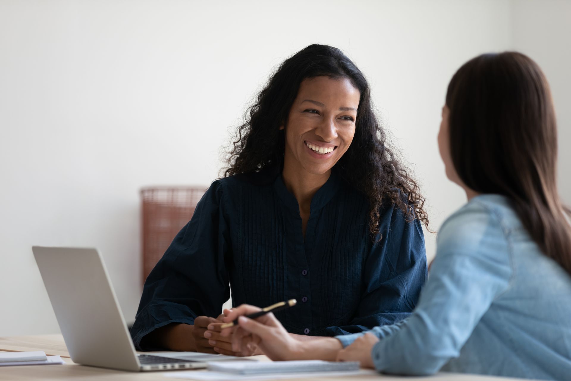Woman smiling while holding pen, speaking to another woman at a desk with laptop.