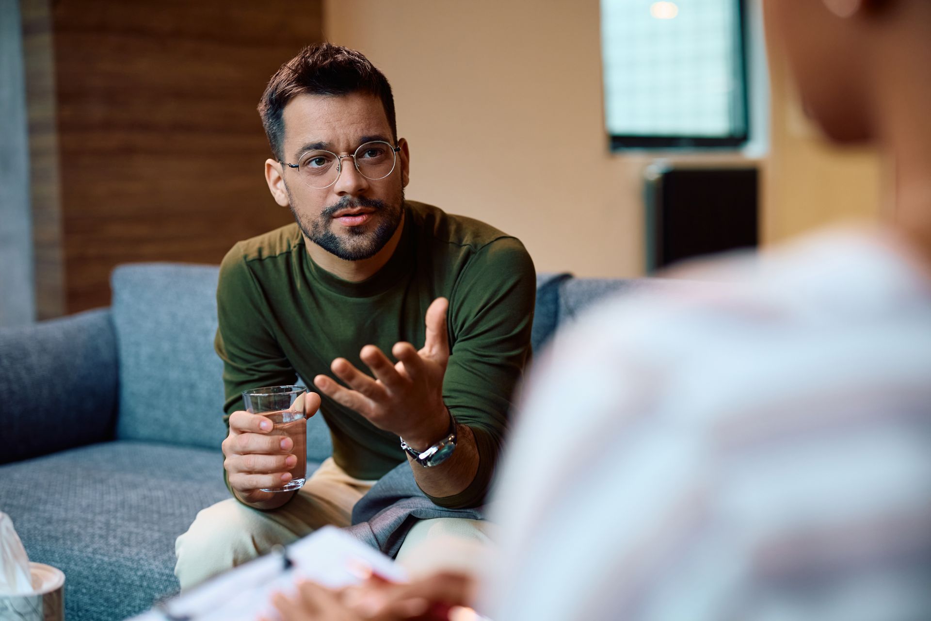 Man in glasses gestures while speaking in therapy session, holding a glass of water.