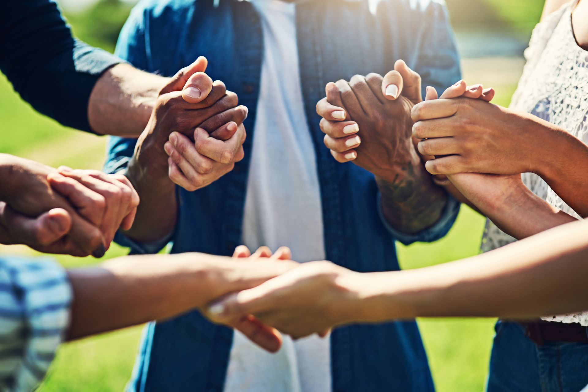 Hands clasped in a circle, diverse skin tones, outdoors, sunlight.