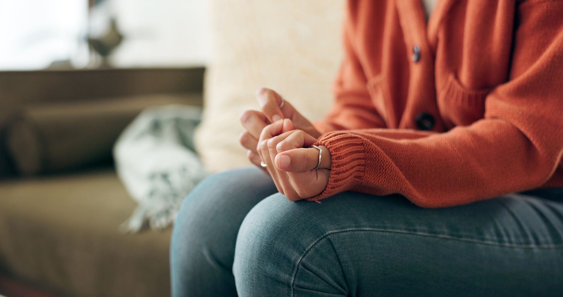 Person seated, clasped hands in lap, wearing orange sweater and jeans; indoor setting.