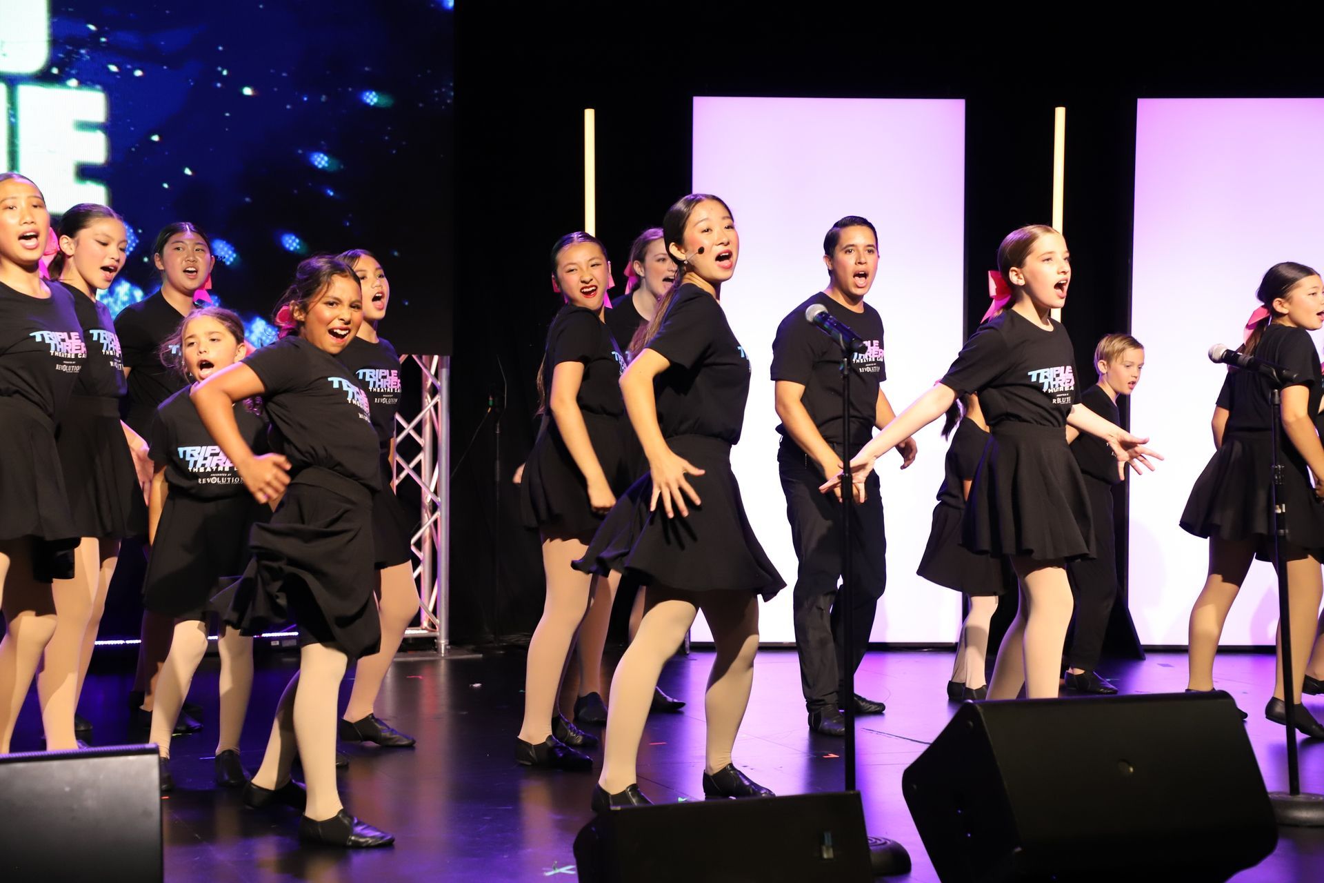 Group of dancers in black outfits performing a synchronized routine on stage under purple lighting
