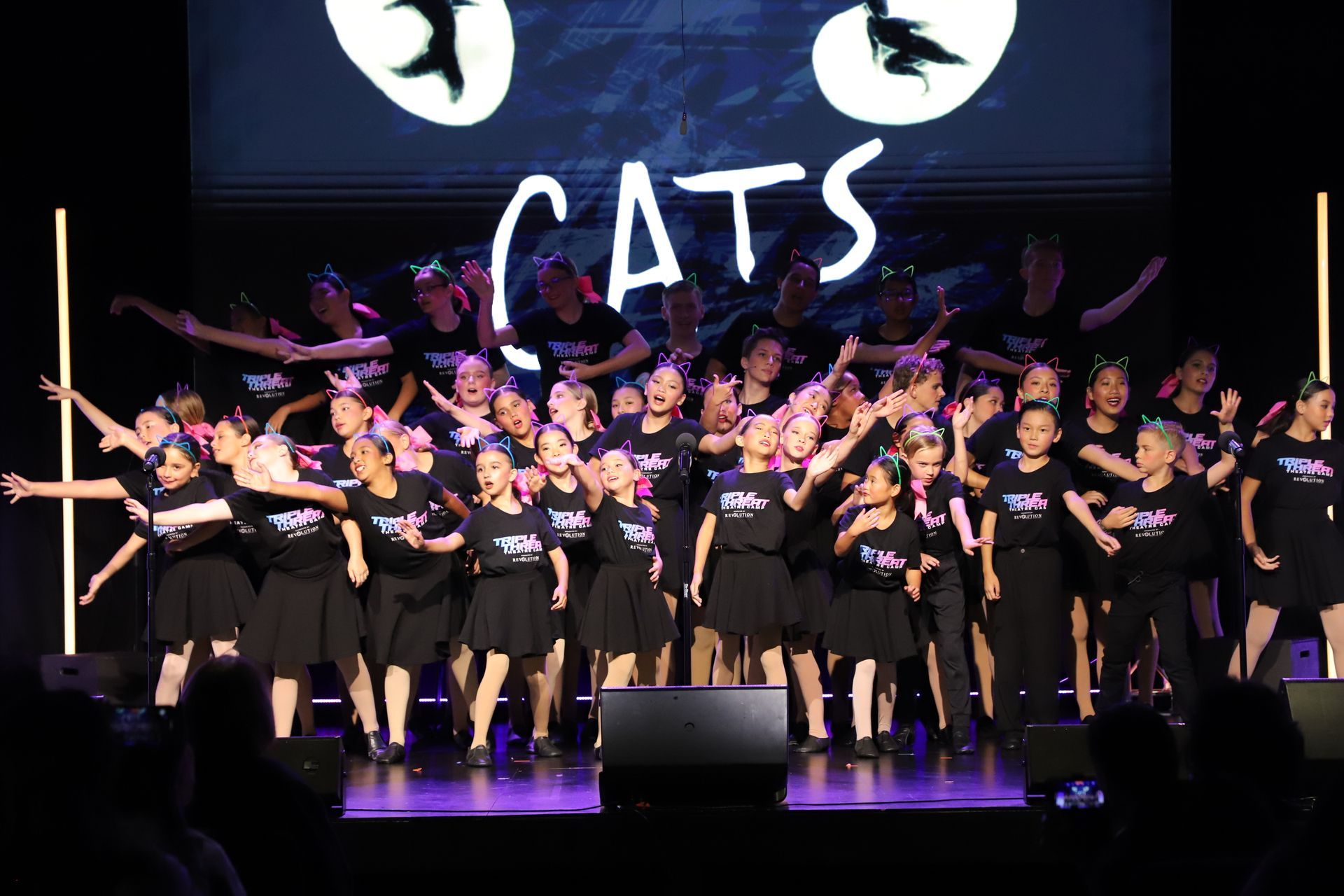 Large group of performers in black costumes dancing on a stage under a CATS sign for a theater showcase.