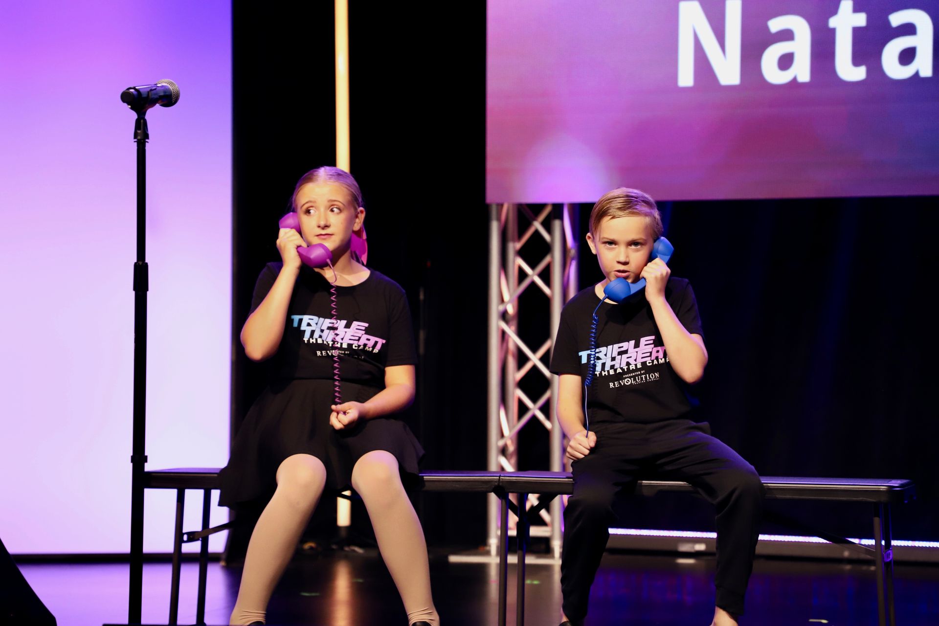 Two children seated on a stage holding microphones and a dance recital setting for a theater showcase.
