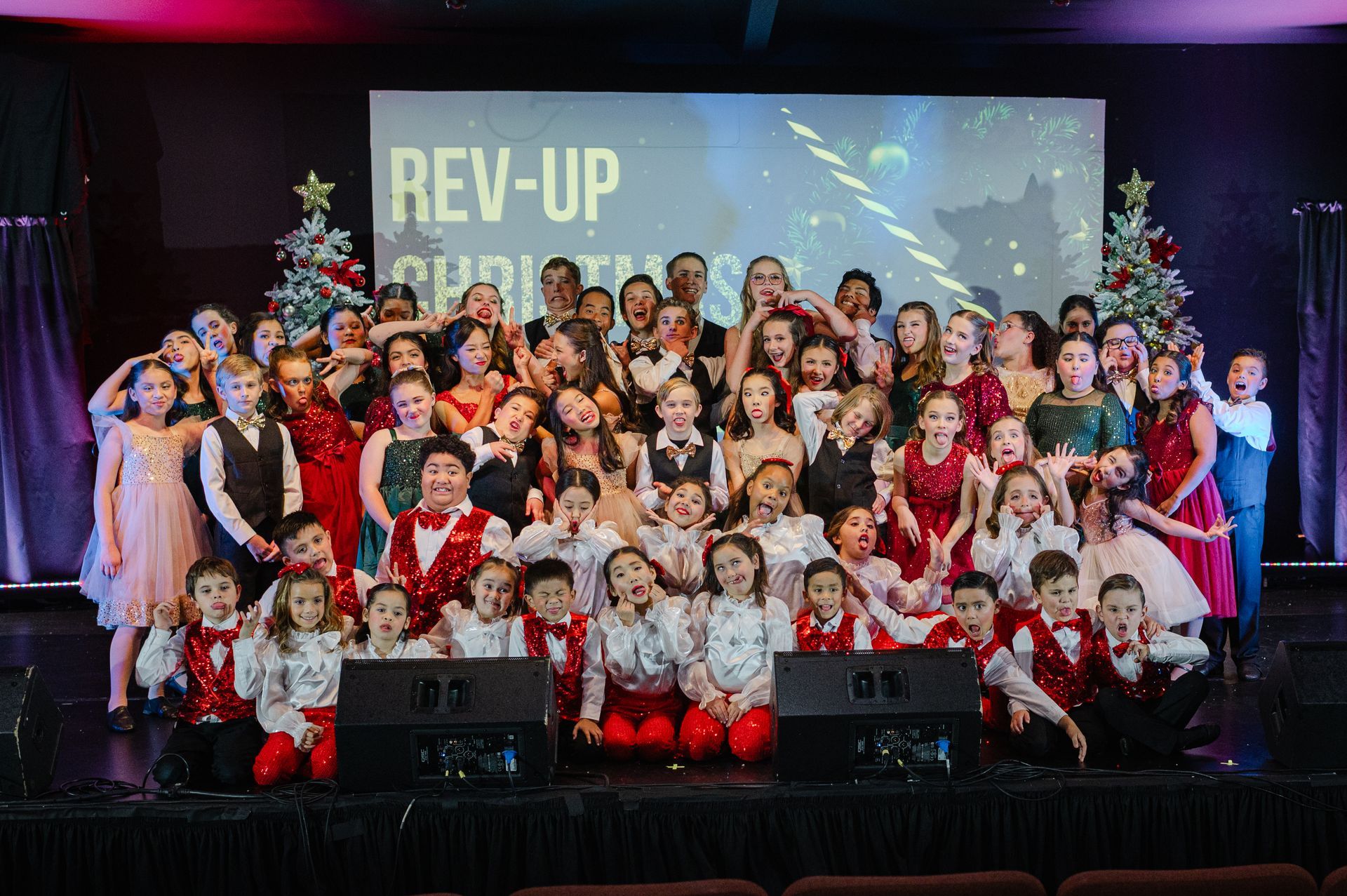 Large group posing on a stage in front of a REV-UP banner, dressed in festive red and white costumes.