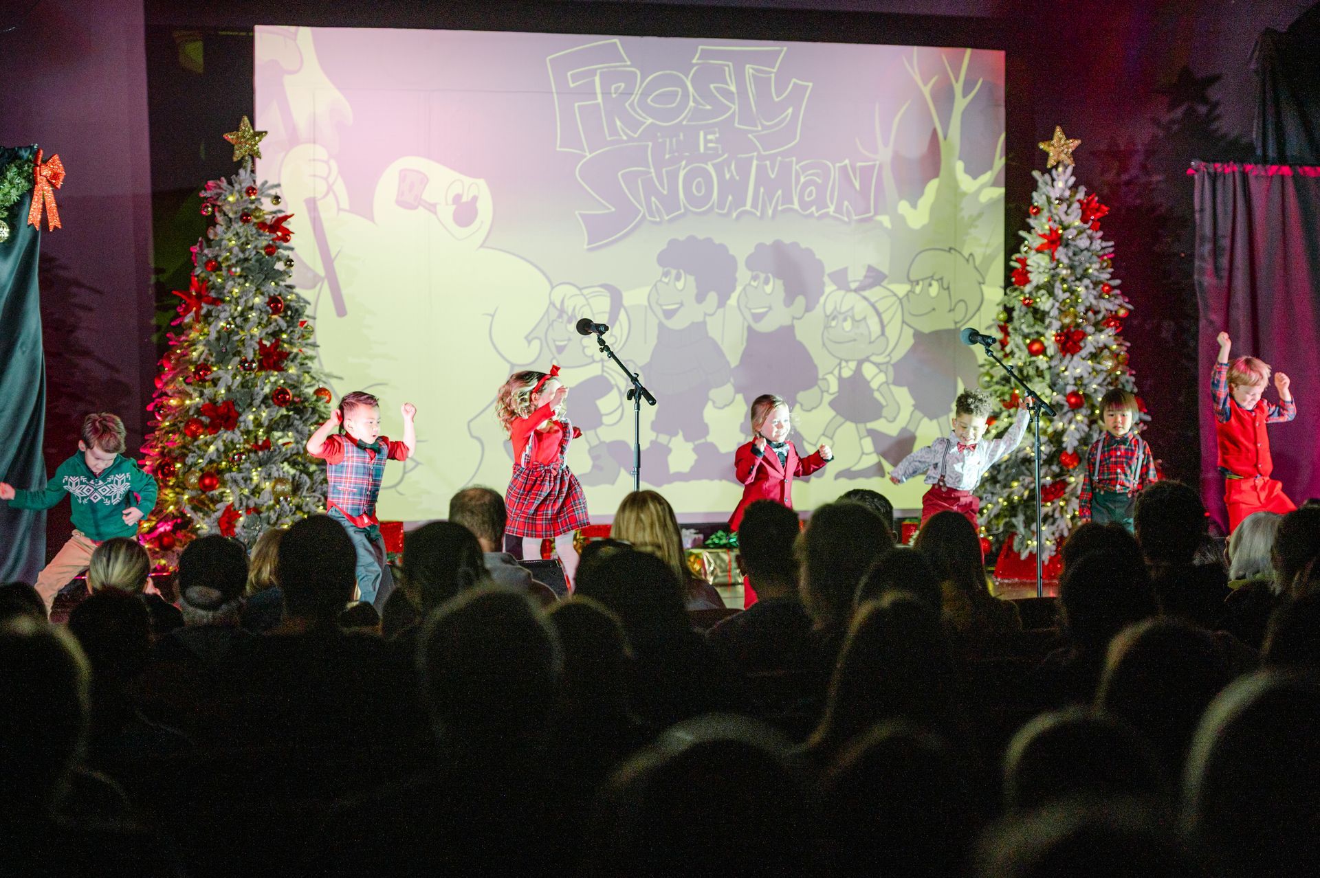 Children perform on a decorated holiday stage with Christmas trees and a snowy winter backdrop.