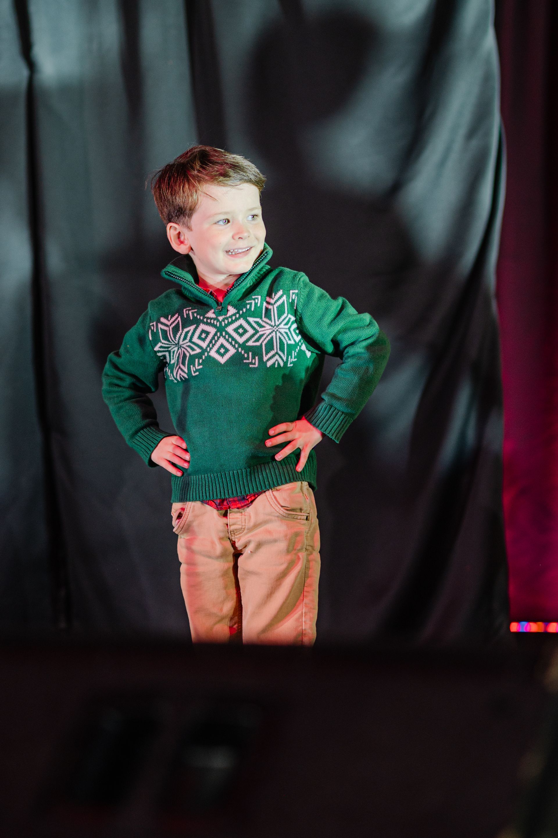 Child posing on a stage in a green sweater and tan pants, hands on hips, smiling against a black backdrop