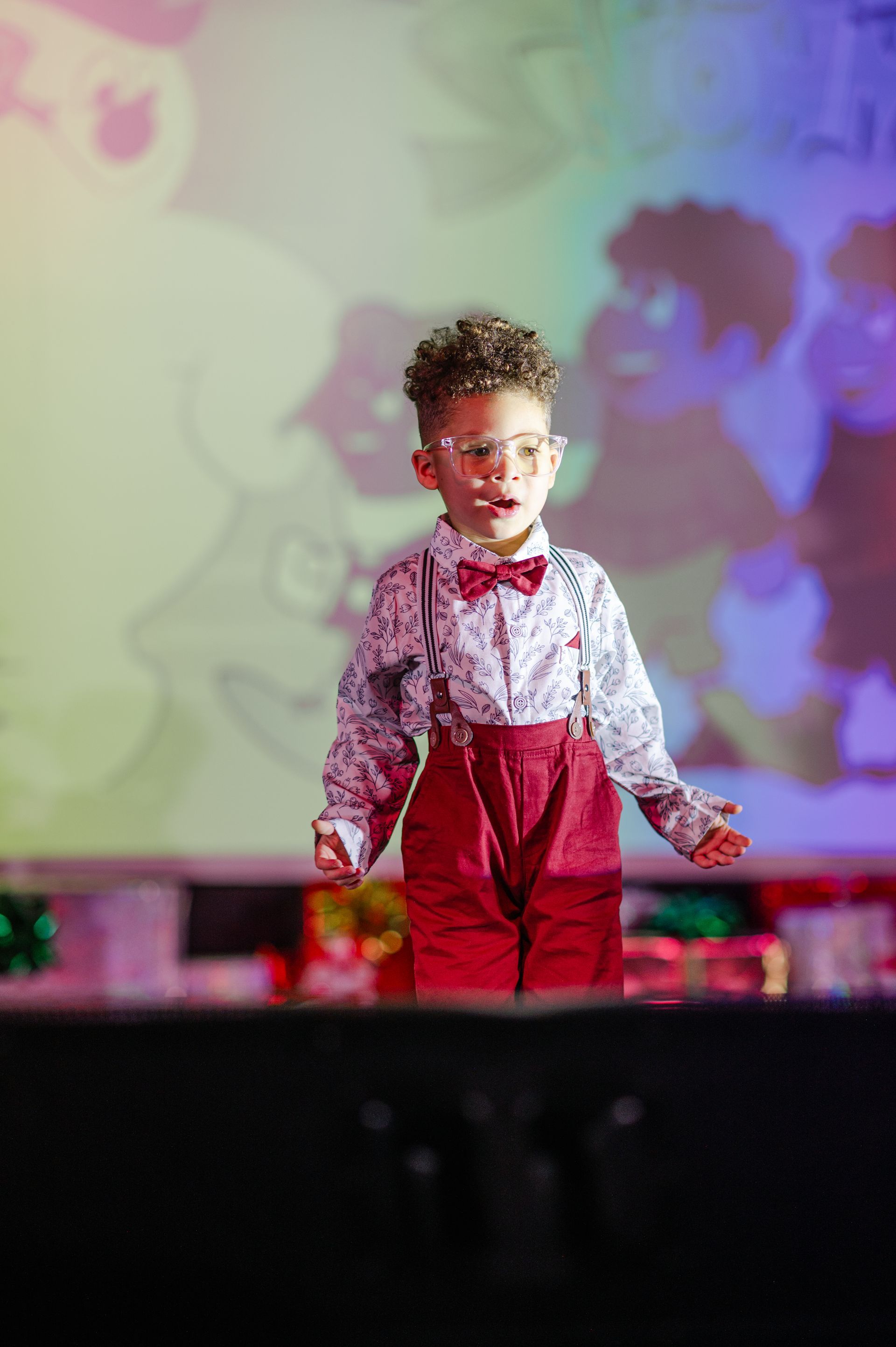 Person in red pants and a patterned shirt standing on a stage with colorful projected lights.