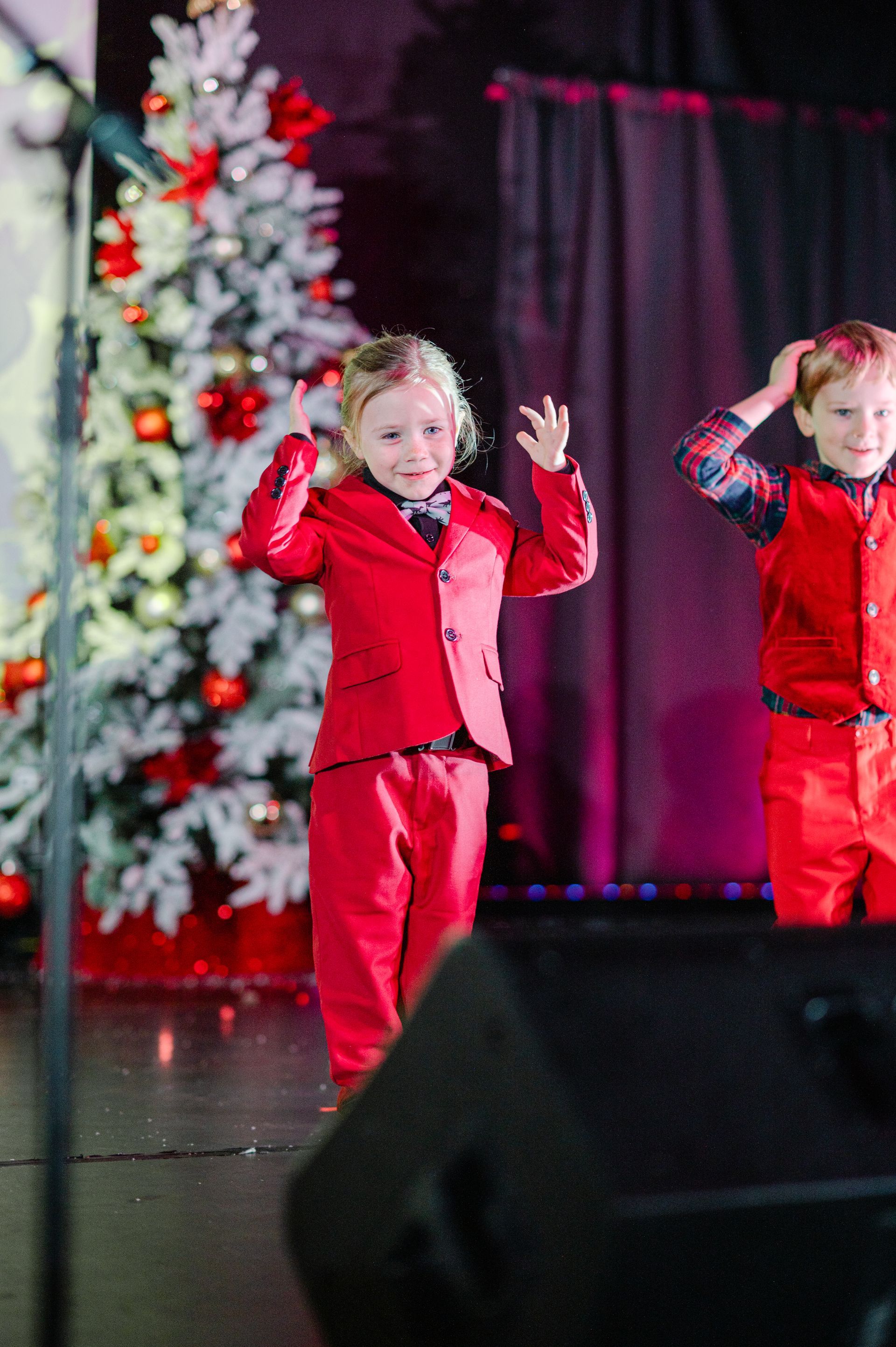 Two children in red outfits dancing onstage beside a decorated Christmas tree.