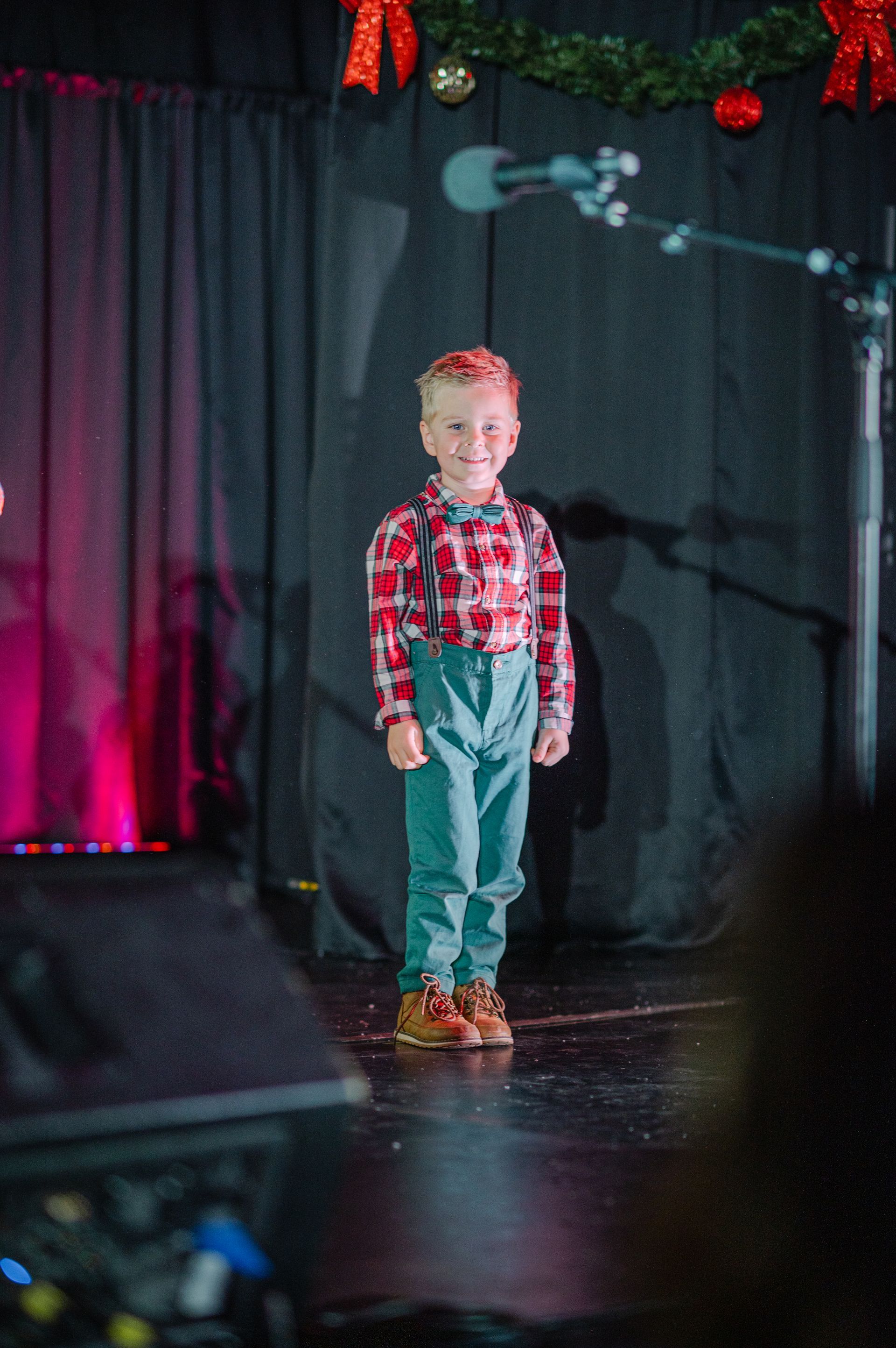 Child in a red plaid shirt and jeans standing on a stage with black curtains and pink lighting.
