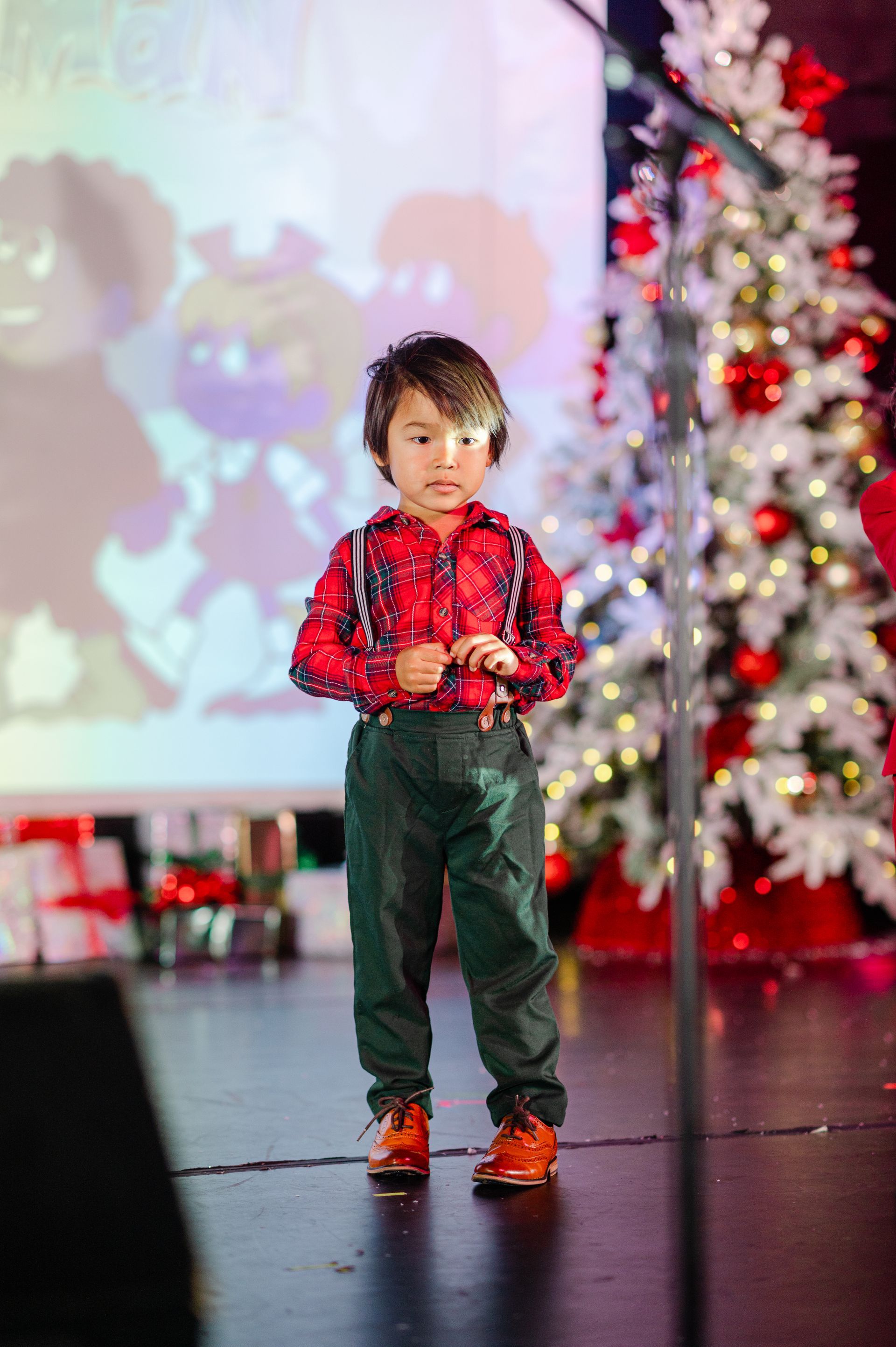 Child in a red plaid shirt and green pants standing on a stage with a decorated Christmas tree behind him.