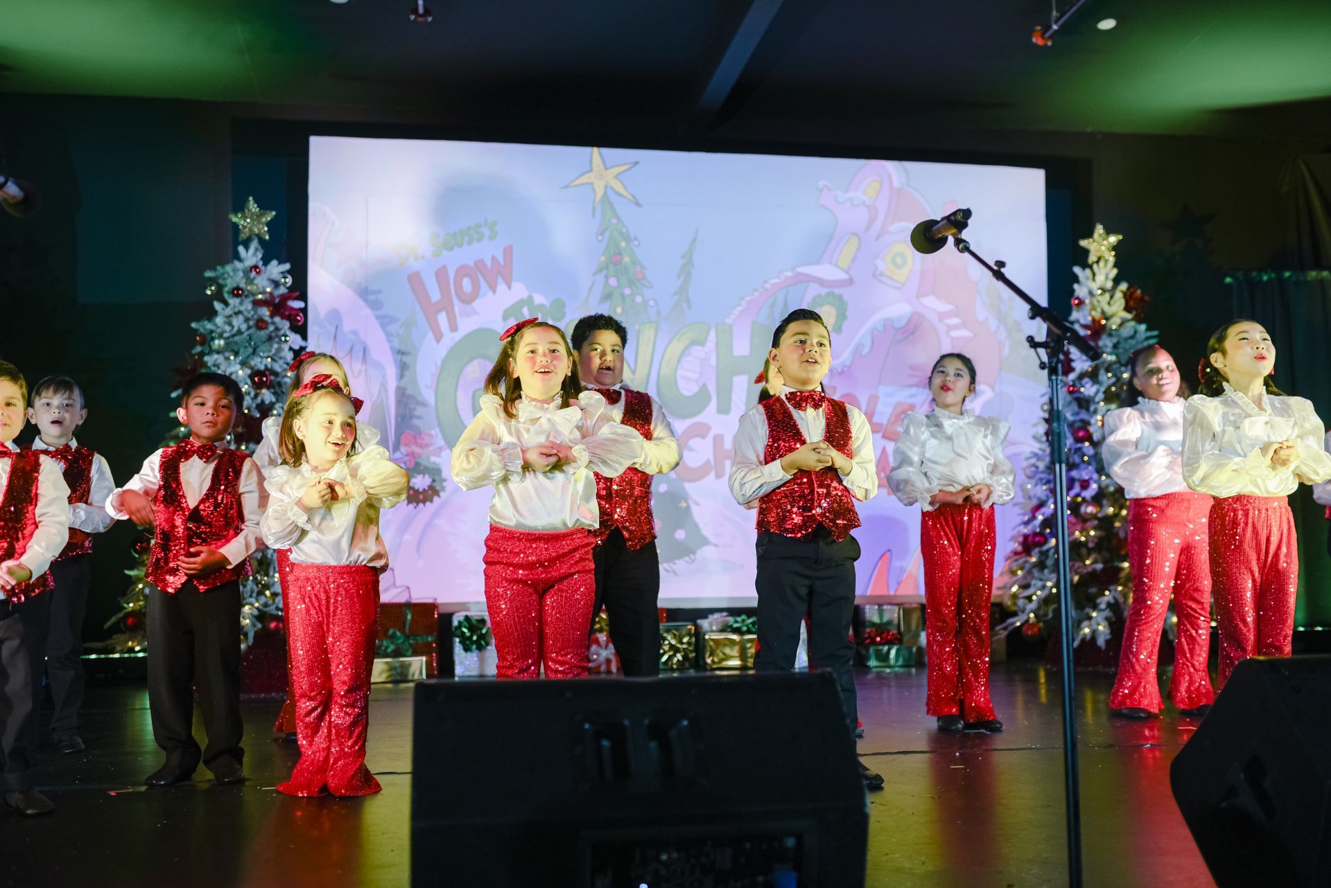 Children in red pants and white tops perform on stage with holiday decorations and a projection screen.