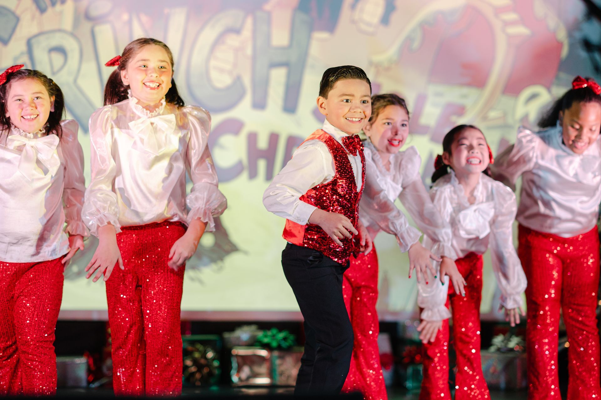 Children performing a dance onstage in red and white costumes, smiling under colorful lights