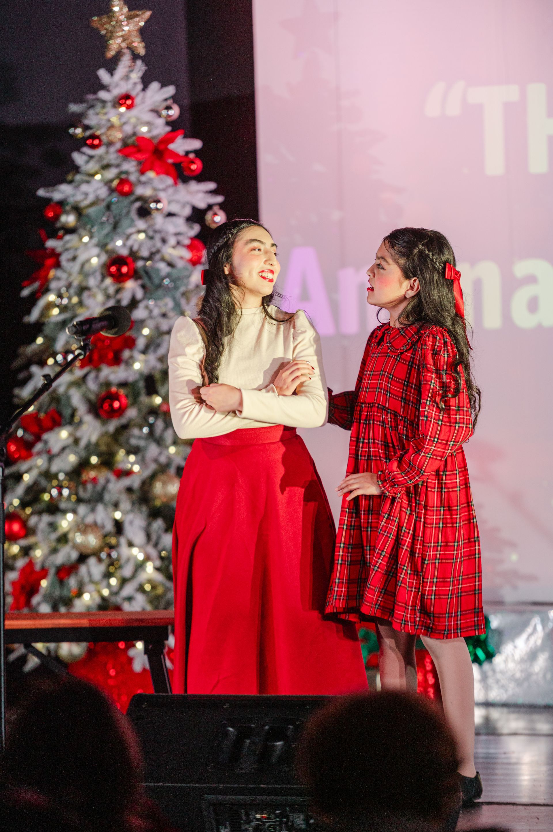 Two children onstage in red outfits beside a decorated Christmas tree, smiling at each other