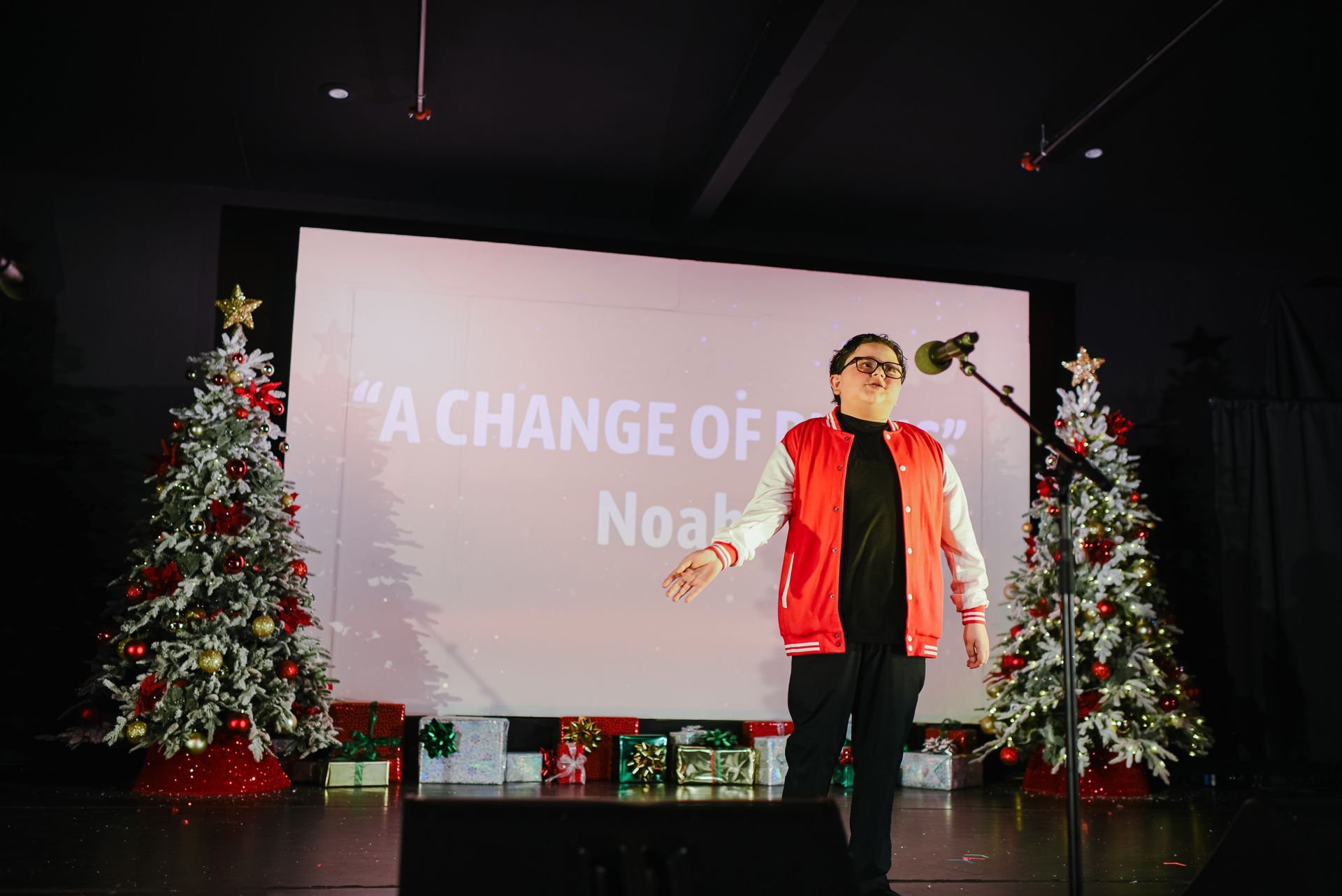 Person on stage beside a microphone, with Christmas trees and a presentation screen behind them.