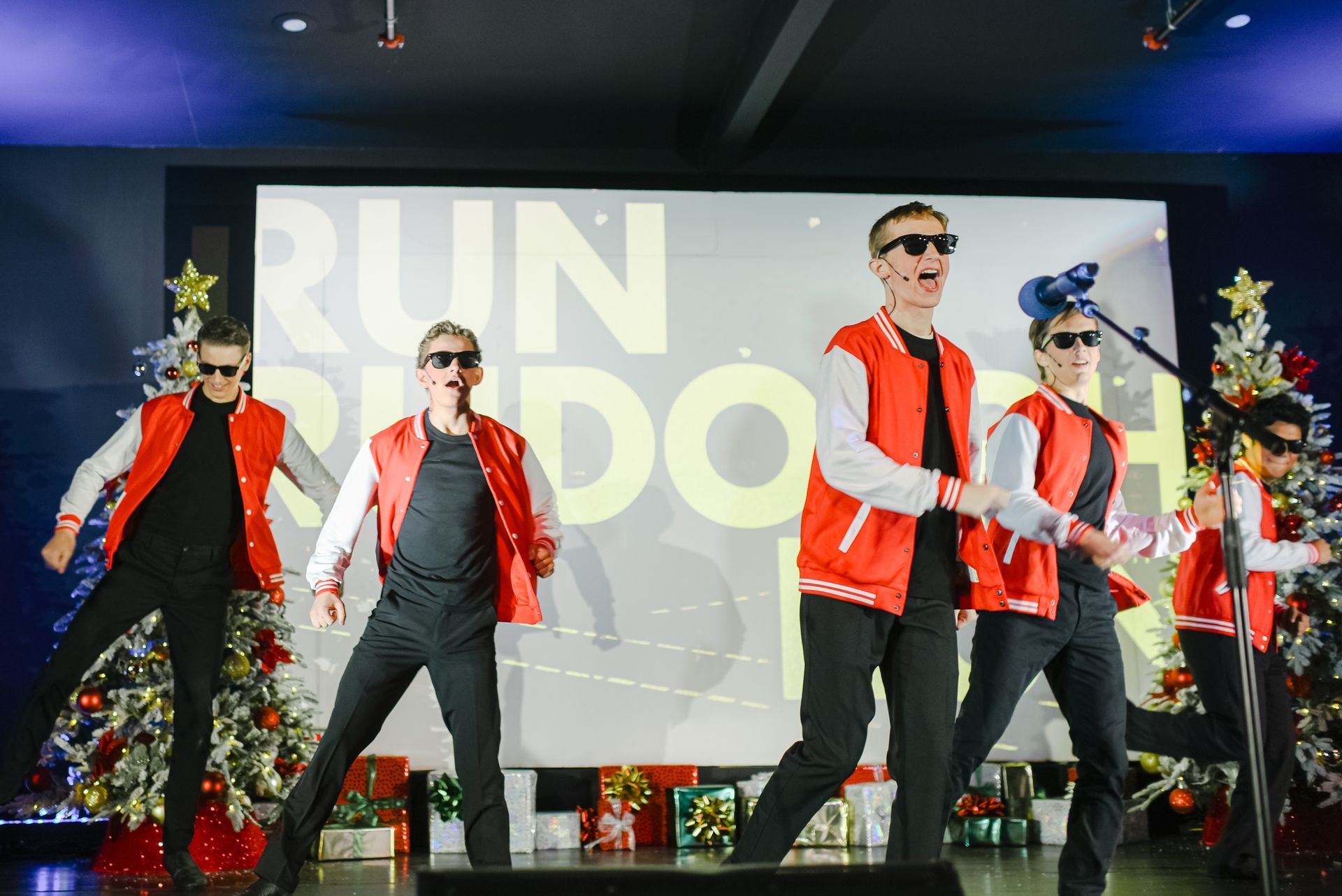 Dancers in red vests and sunglasses perform on stage with a “RUN” backdrop and Christmas trees