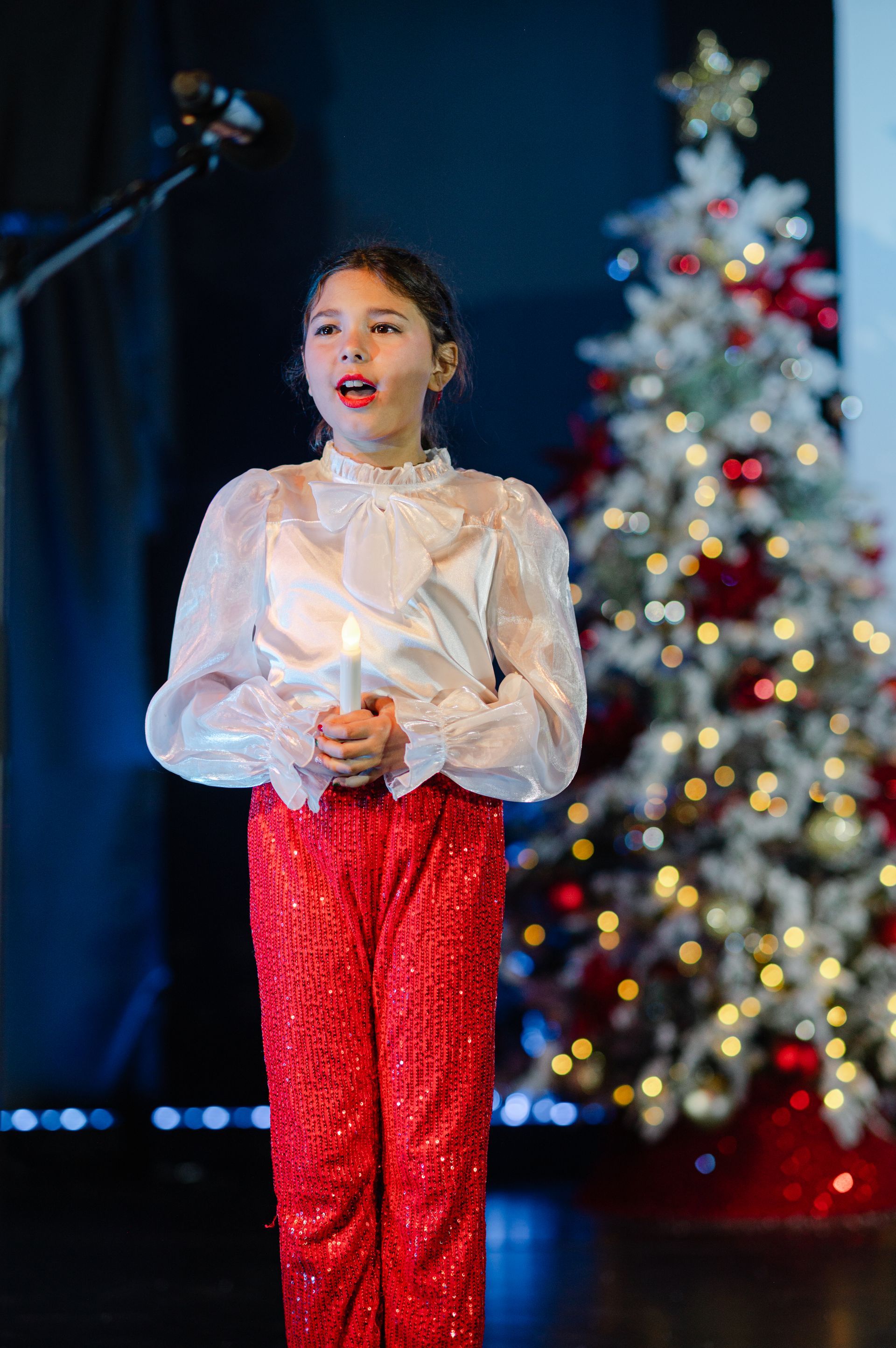 Young performer in a white blouse and red pants singing on stage beside a lit Christmas tree