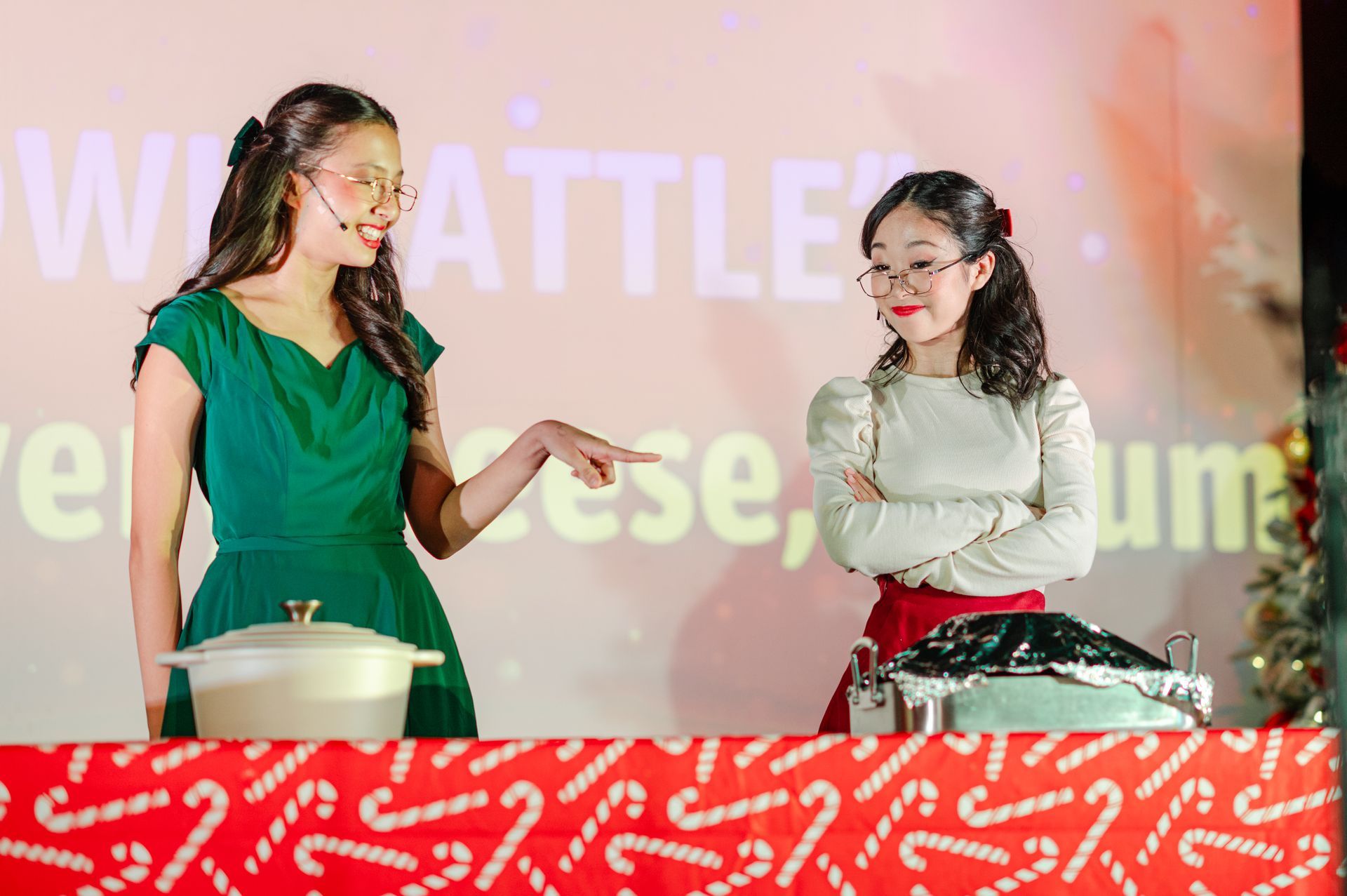 Two women on stage in green and white dresses, smiling and gesturing behind a red-covered table