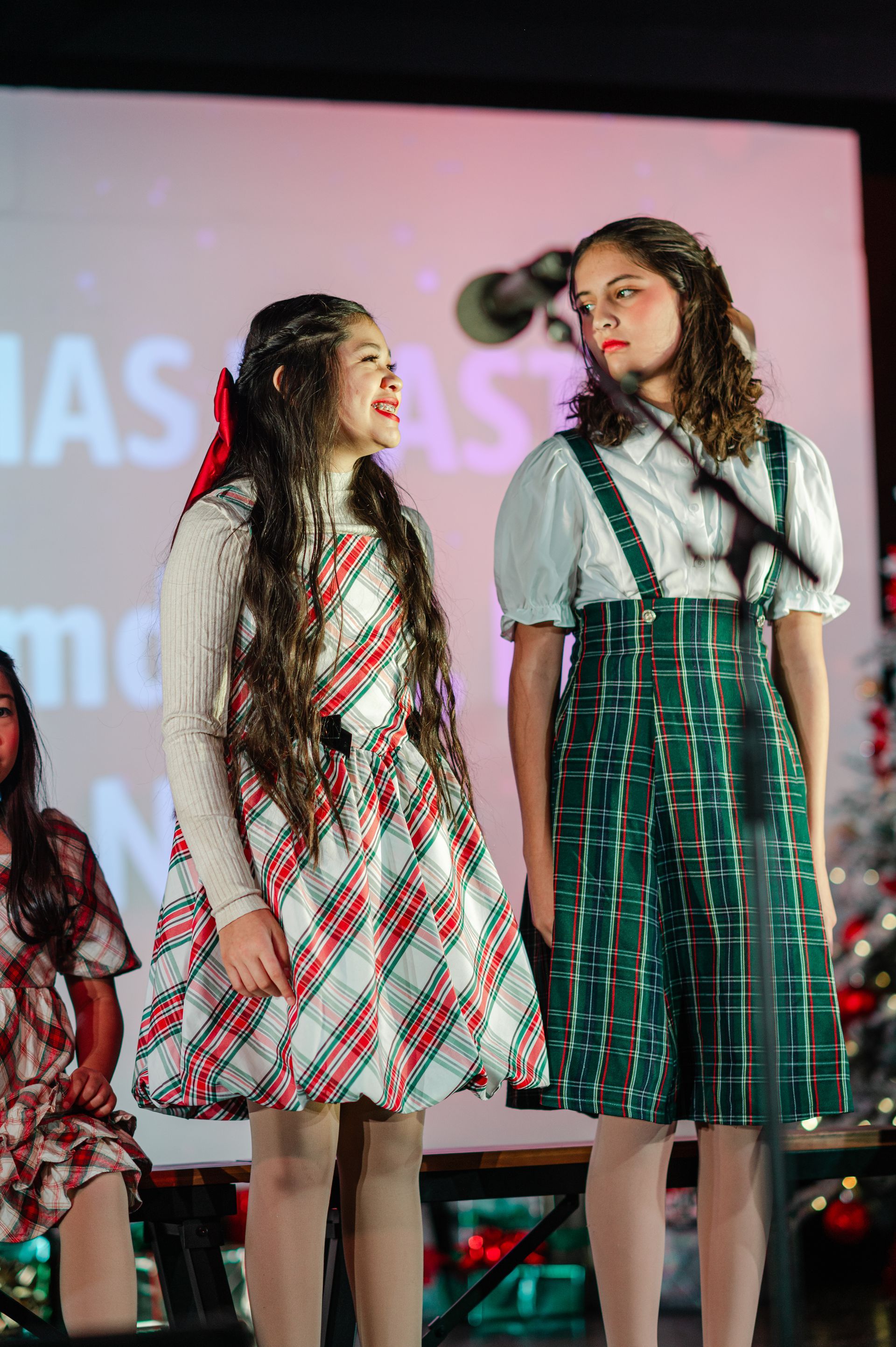 Two performers in patterned dresses standing on a stage, facing each other before a lit screen