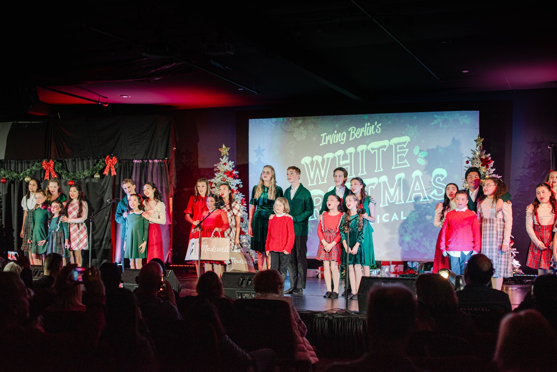 Children performing on a decorated stage with Christmas trees and a holiday backdrop, audience silhouetted in front.