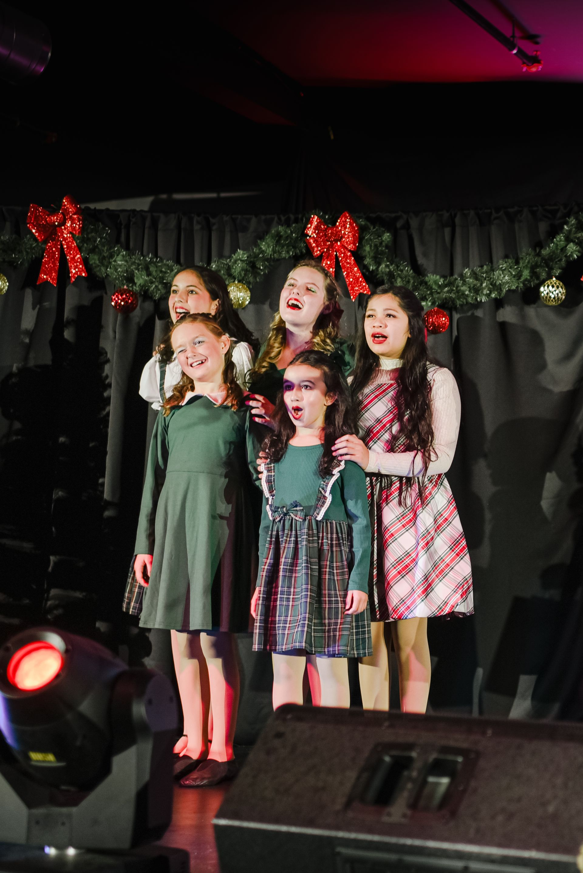Three girls singing on a decorated stage with holiday lights and red bows
