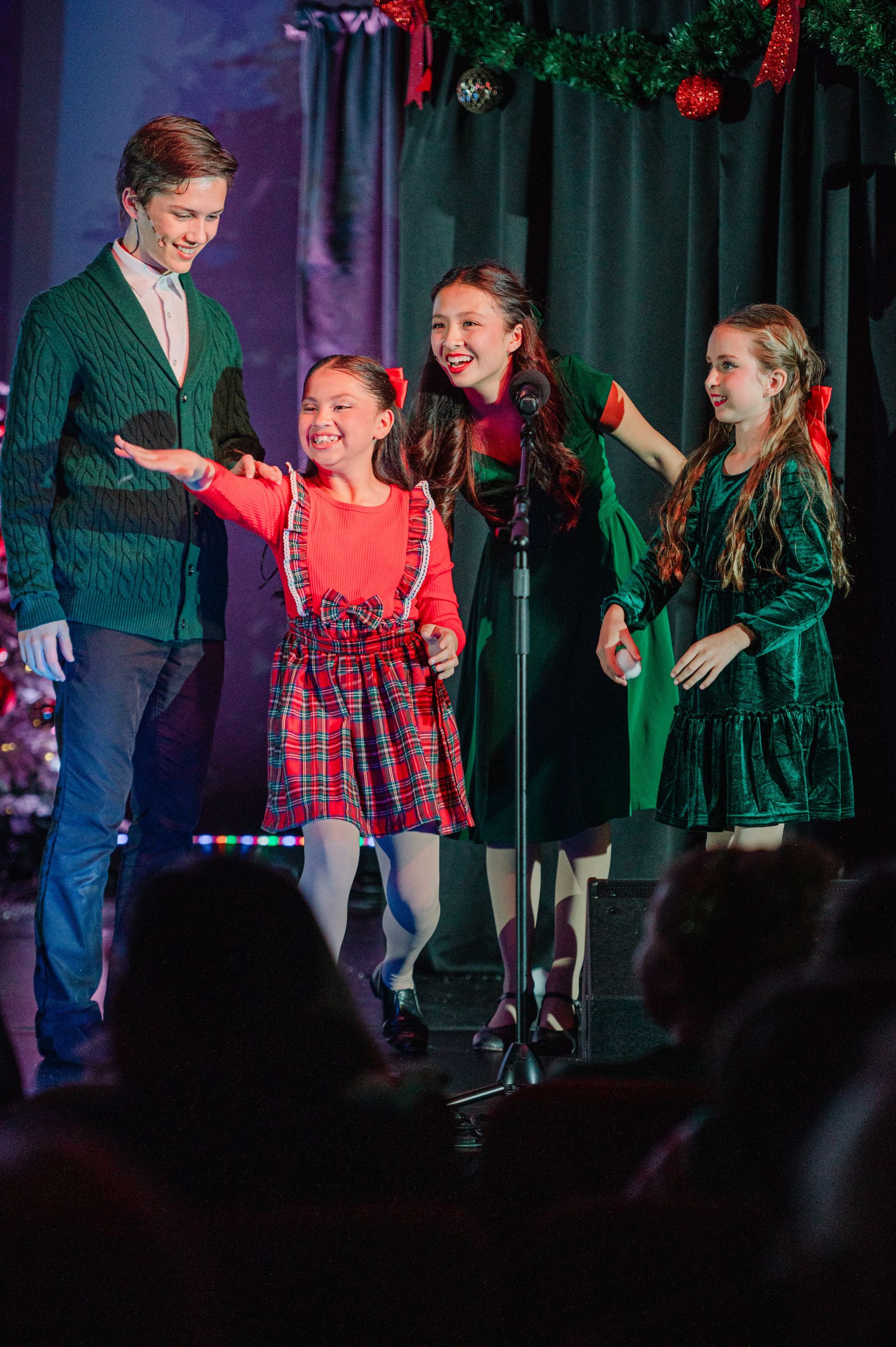 Four people on stage singing, with two children in green and red holiday outfits under festive lights.