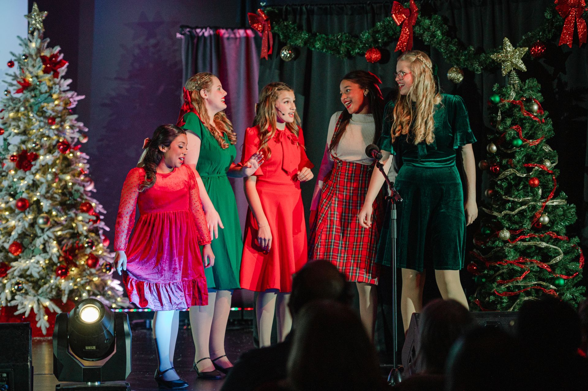 Five women in festive dresses sing on a decorated holiday stage with Christmas trees and lights.