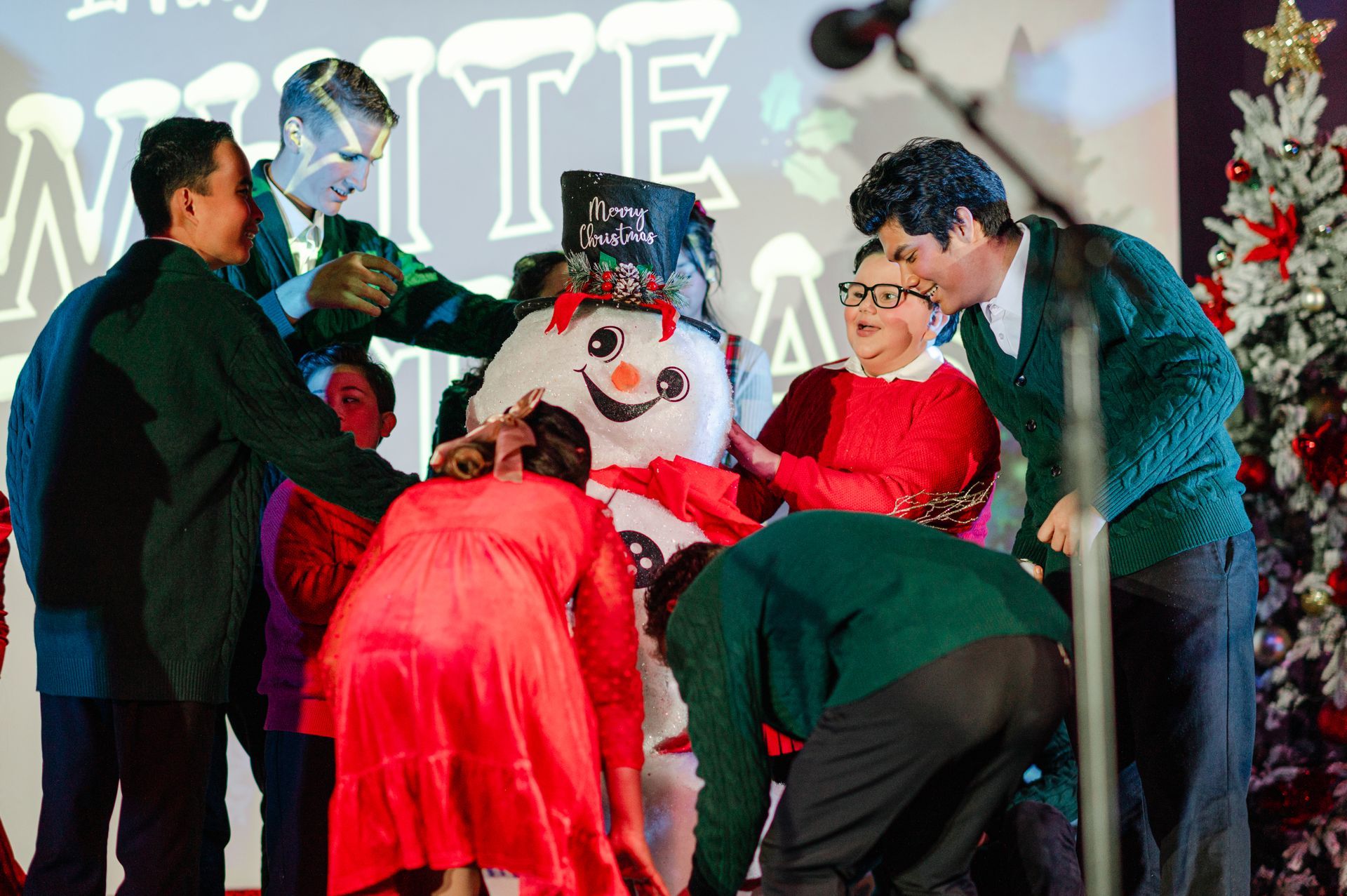 People gathered around a giant snowman on a decorated stage with a Christmas tree.