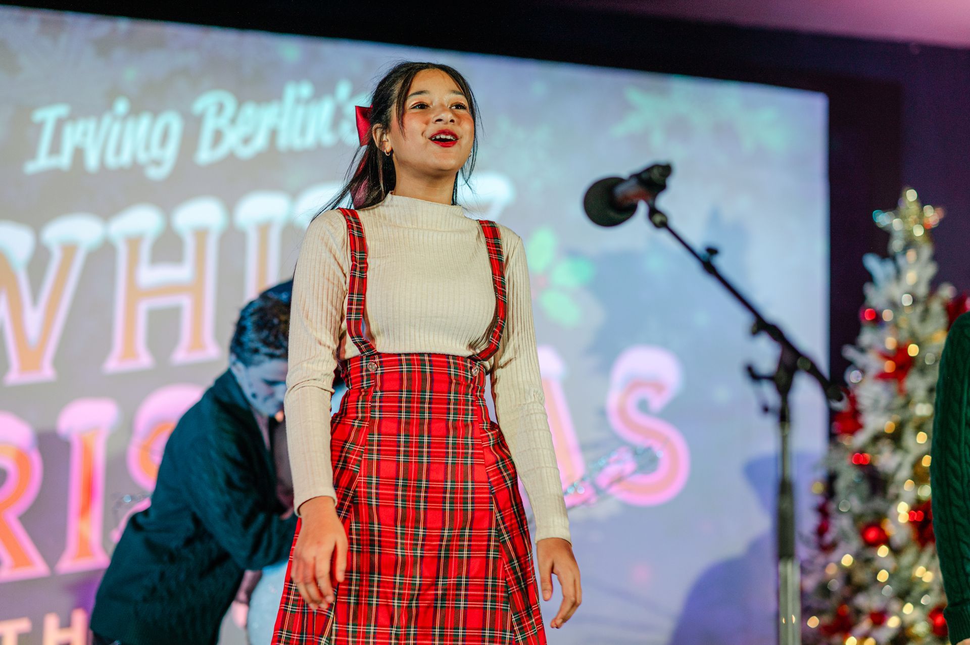 Woman in a red plaid dress standing onstage with a microphone and festive lights in the background