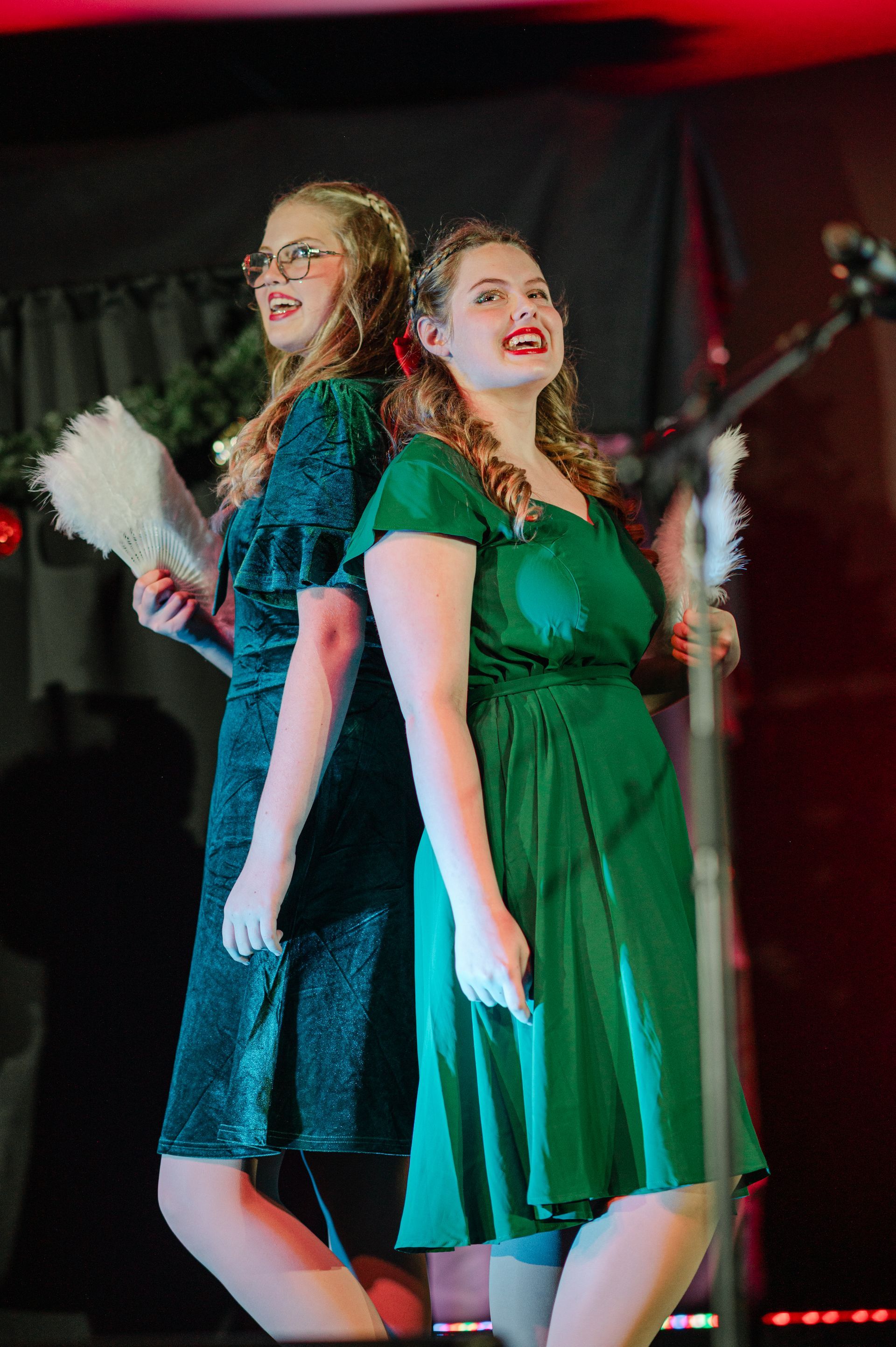 Two performers in green dresses stand back-to-back on a stage, smiling under red lighting.