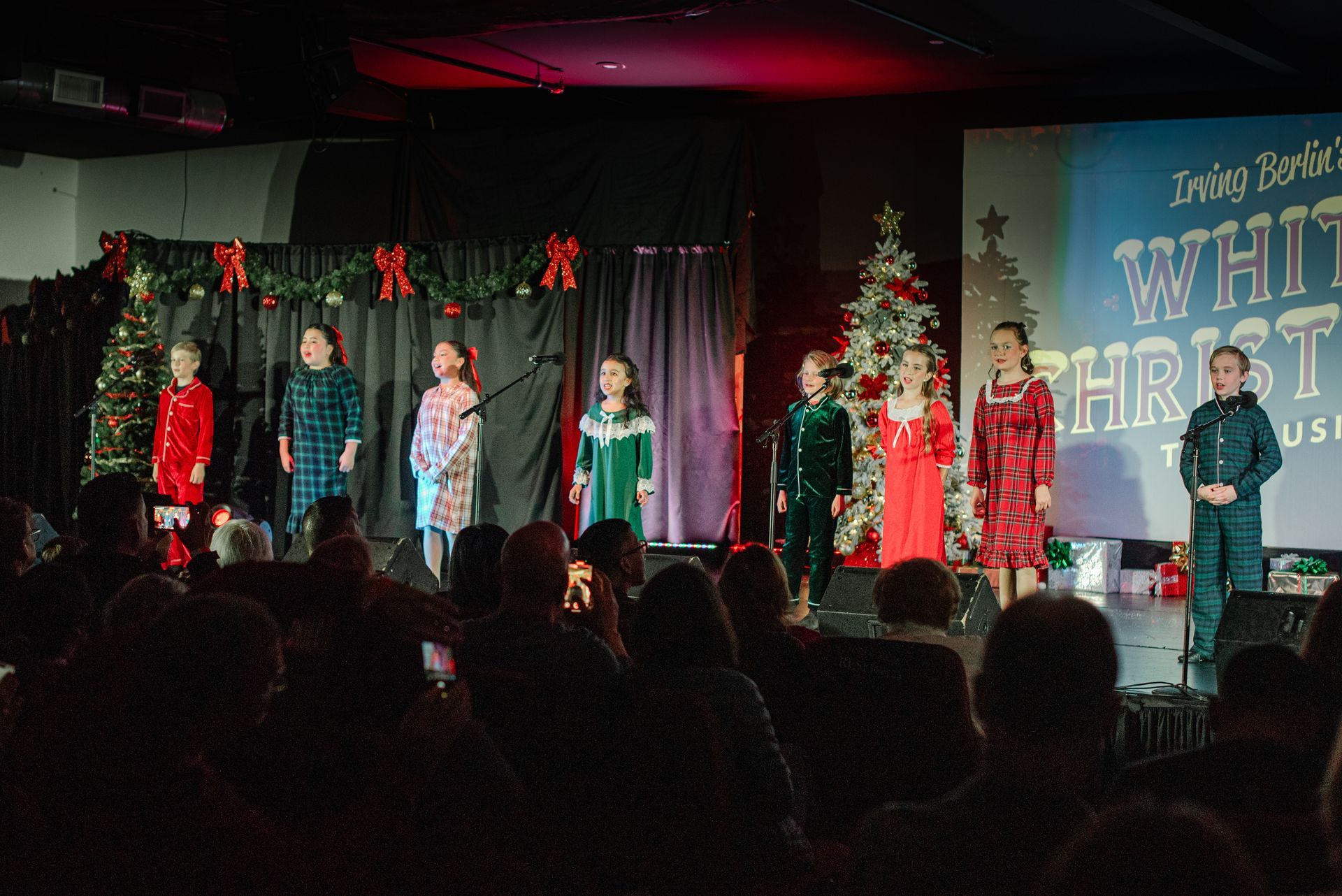 Children singing on a decorated stage during a holiday concert, with an audience watching.